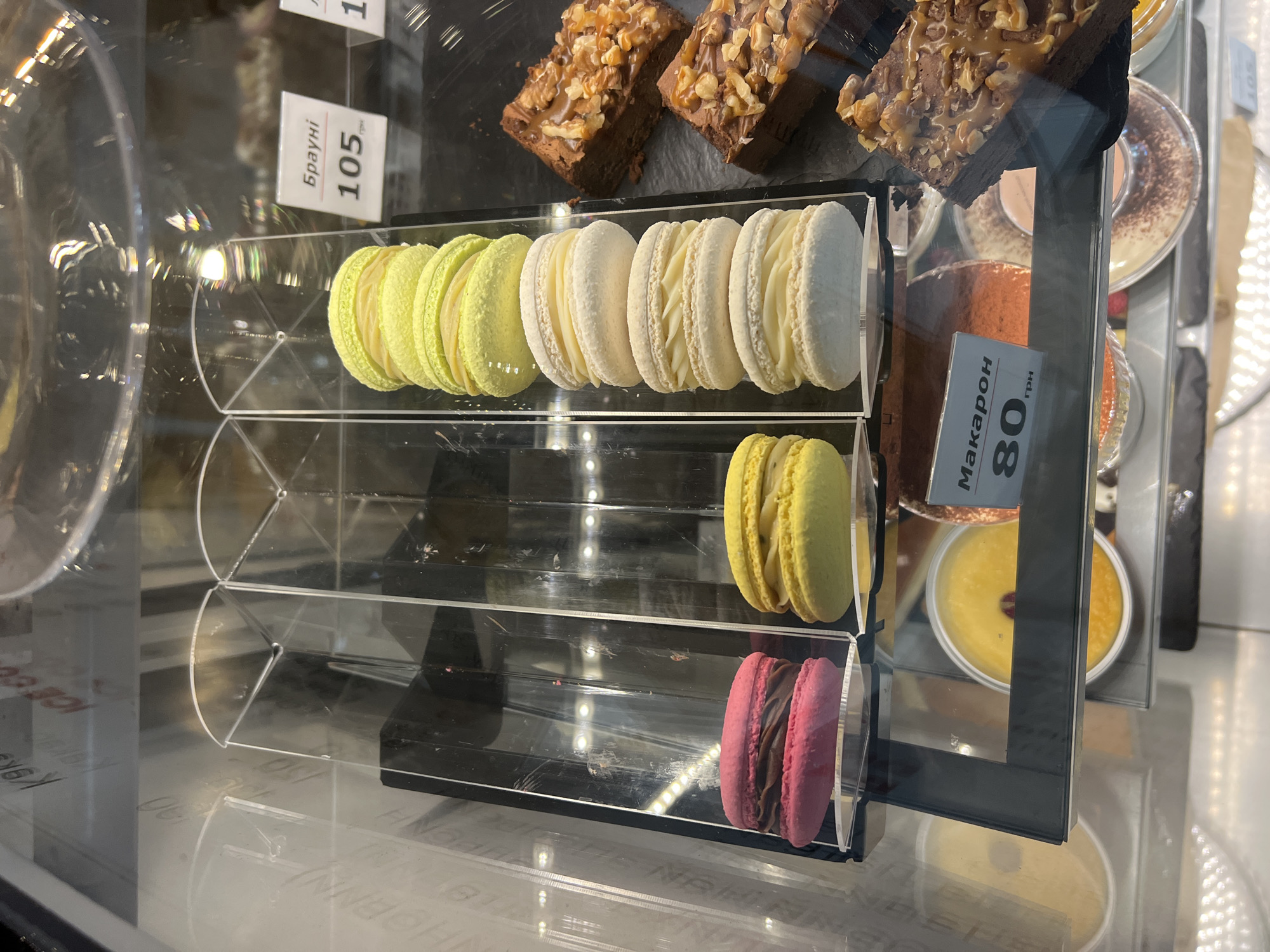 This is a medium-angle, eye-level shot of a refrigerated display case in a food establishment, likely a bakery or cafe, located in Fontanka, Ukraine. The display case is made of glass and metal and is illuminated from above and behind.
Front and center, there are several macarons stacked vertically in clear plastic compartments. The top compartment contains a stack of three pale green macarons, each filled with a lighter green cream. Below this, another compartment holds four white macarons, also filled with a cream. To the left, a pink macaron sits by itself in a compartment. To its right, a single yellow macaron is visible, filled with a creamy substance.
To the right of the macarons, slices of brownies are arranged on a dark surface. The brownies are topped with chopped nuts and a caramel drizzle.
Below the macarons, a price tag is visible with Cyrillic text that appears to say "Макарон" and "80 грн", indicating a price of 80 Ukrainian Hryvnia for macarons.
In the background, more food items are visible in the display case, including what appear to be desserts in small cups or bowls, some with orange and yellow fillings. The overall environment suggests a commercial food service setting, with a focus on pastries and desserts.
