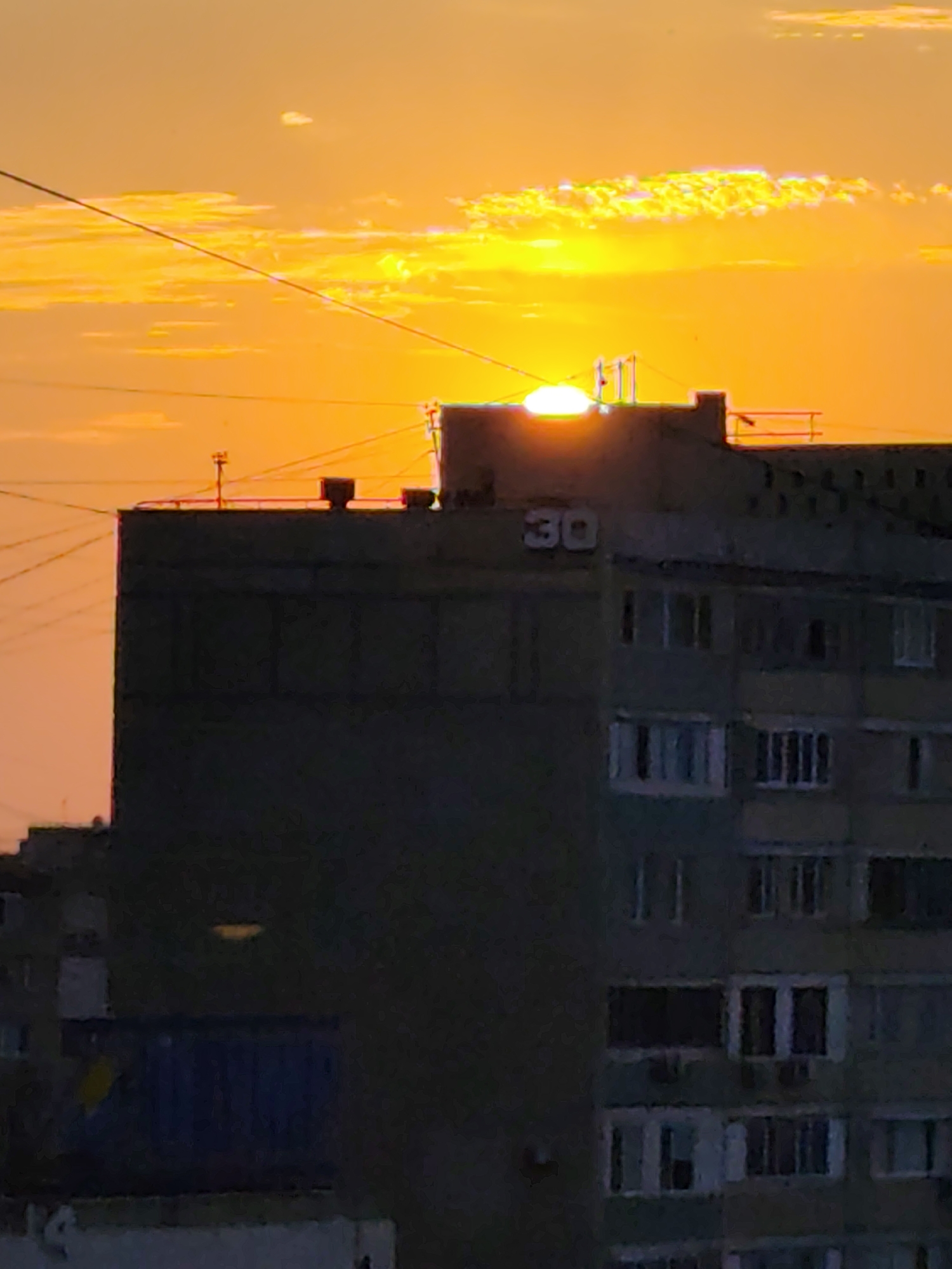 A bright orange sunset illuminates the sky above a silhouetted apartment building in Aktau, Kazakhstan. The sun, a glowing orb, is partially obscured by the roofline of the building. Several wires crisscross the sky, with a few visible antenna-like structures on the building's roof. The building itself is dark and appears to be an older, concrete structure, with many windows visible. The number "30" is painted in white on the side of the building near the top. In the lower left corner, a dark blue container with a yellow marking is partially visible. The overall scene captures a typical urban sunset in Aktau.