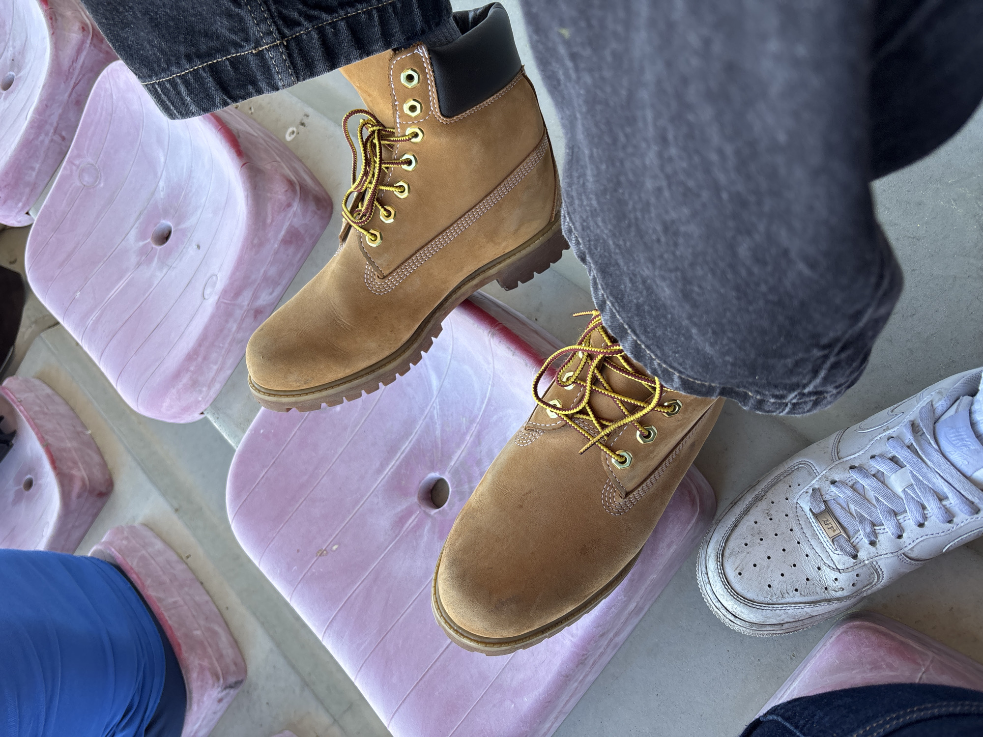 The image presents a low-angle, eye-level shot of a person seated in a stadium setting. The individual's feet, clad in two different types of footwear, are prominently displayed. One foot wears a tan-colored Timberland-style work boot with yellow and burgundy laces, while the other sports a white Nike Air Force 1 sneaker. The person is wearing dark denim jeans.
The seating appears to be made of molded pink plastic, with several rows visible in the background. The setting suggests an outdoor stadium or arena.
While the exact location within Valpaços, Portugal cannot be determined solely from the image, the stadium seating suggests a sports facility or a similar public gathering space within the city. No other individuals or specific landmarks are visible in the frame to pinpoint the location further.