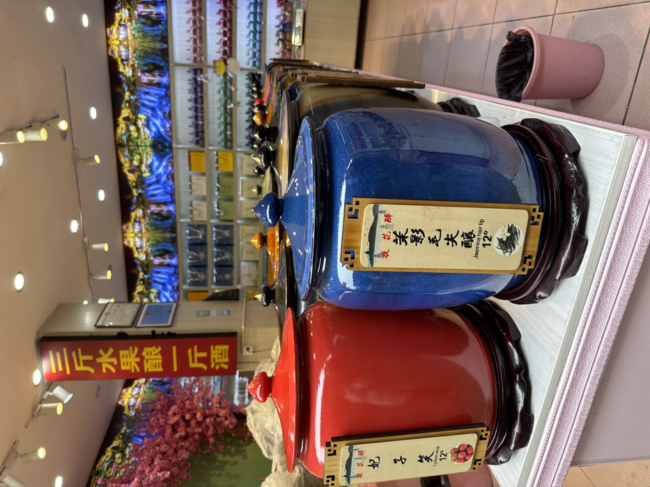This image depicts the interior of a retail shop in Furong, China. In the foreground, a display counter with a light pink, textured trim presents two large, lidded ceramic jars on dark wooden stands. The jar on the left is glossy red with a matching conical lid, displaying a rectangular wooden sign that reads "妃子笑" (Feizi Xiao), "Lychee wine," and "12°," alongside an image of lychees. The jar on the right is speckled blue with a matching conical lid, displaying a rectangular wooden sign that reads "茉莉毛尖" (Moli Maojian), "Jasmine hair tip," and "12°," alongside an image of jasmine tea leaves. Both signs also feature the characters "夜花醉" (Ye Huazui). A row of additional similar lidded jars with various colored lids (black, orange, blue, brown) extends along the counter into the background.
The shop's left wall is adorned with artificial pink cherry blossoms. The rear wall features a large, illuminated mural depicting a vibrant landscape with waterfalls. Below this mural, shelves are stocked with numerous bottles and packaged goods. A vertical red sign with yellow Chinese characters on the left reads "三斤水果酿一斤酒" (Three catties of fruit make one catty of wine). Overhead, multiple recessed spotlights illuminate the scene. A small, pink ribbed plastic trash can containing a black liner is visible on the tiled floor to the right of the blue jar. No individuals are present within the frame.