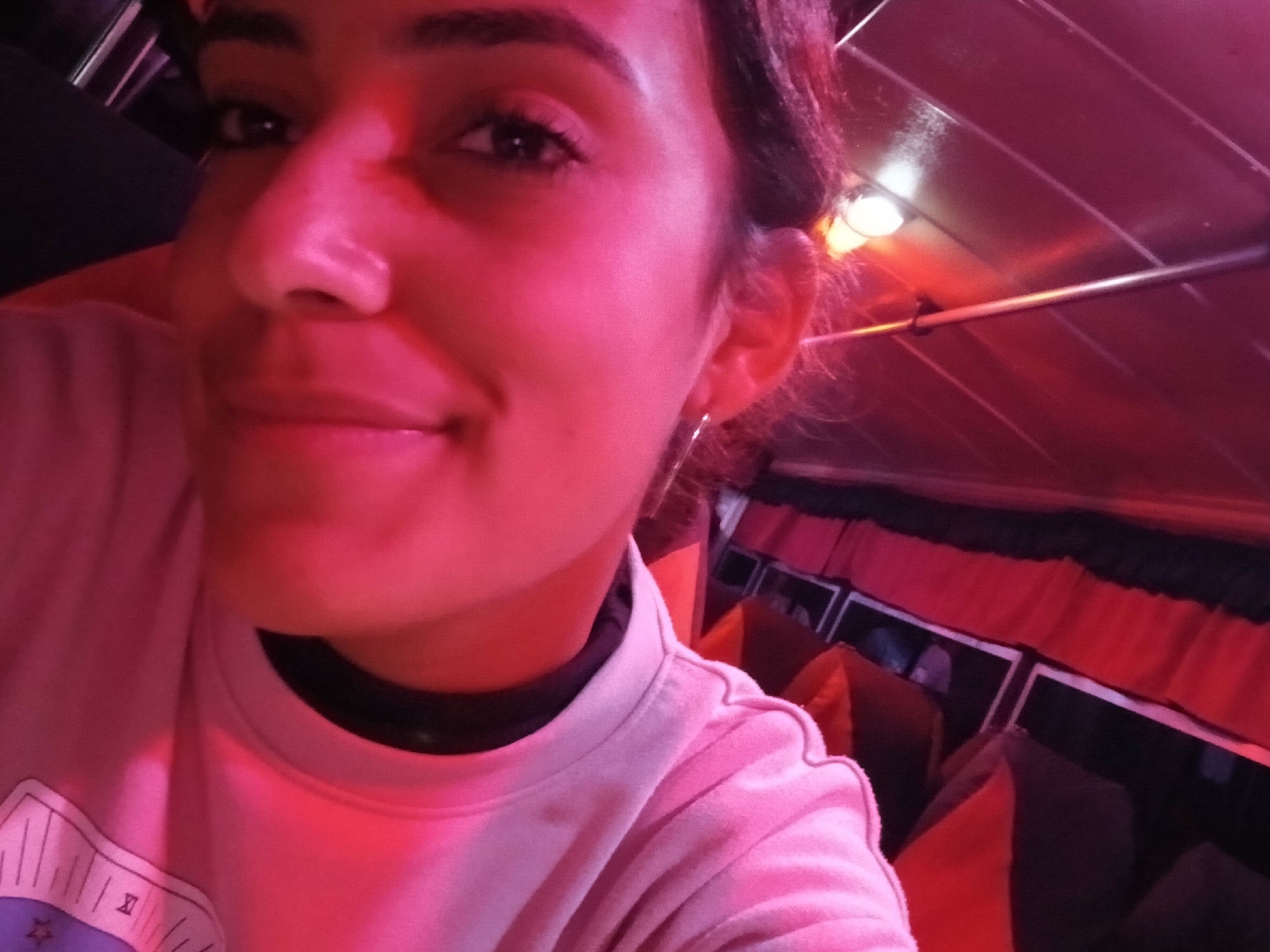 A close-up, angled selfie shows a young woman with dark hair and makeup. She is wearing a pink sweatshirt. The lighting is a strong red hue, likely from ambient lighting. The background appears to be the interior of a dimly lit establishment, possibly a bar or restaurant, with red and black fabric draped along the ceiling. The image does not contain any information about the city of Altagracia, Venezuela.