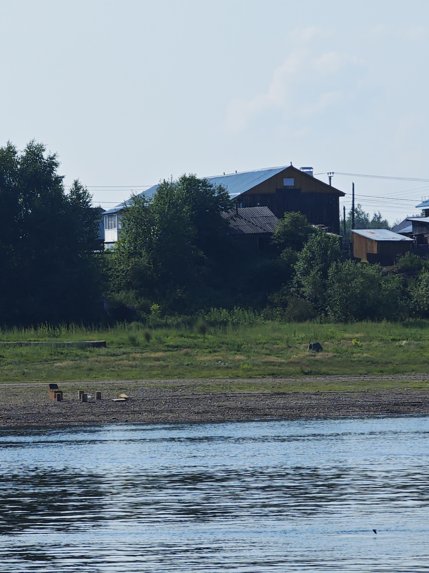 The image depicts a waterfront scene in Verkhnepashino, Russia, on a clear day. In the foreground, a body of blue water with ripples covers the lower portion of the frame. Along the shore, a narrow strip of grey pebbles is visible, transitioning to green grass and foliage further inland. Several wooden structures, appearing to be benches or small tables, are scattered on the pebble area.
Behind the waterfront, a slope is covered with lush green vegetation and trees. Dominating the middle ground are several buildings with pitched roofs. One large, dark-colored building with a yellow-brown upper section and a blue roof is prominent, featuring a chimney and a small window. To its left, a smaller building with a corrugated roof is visible. To the right, a smaller, wooden outbuilding with a corrugated metal roof stands. More trees and vegetation surround these structures, with power lines visible in the background. The sky above is pale blue with a few wispy clouds. There are no visible people in the scene.