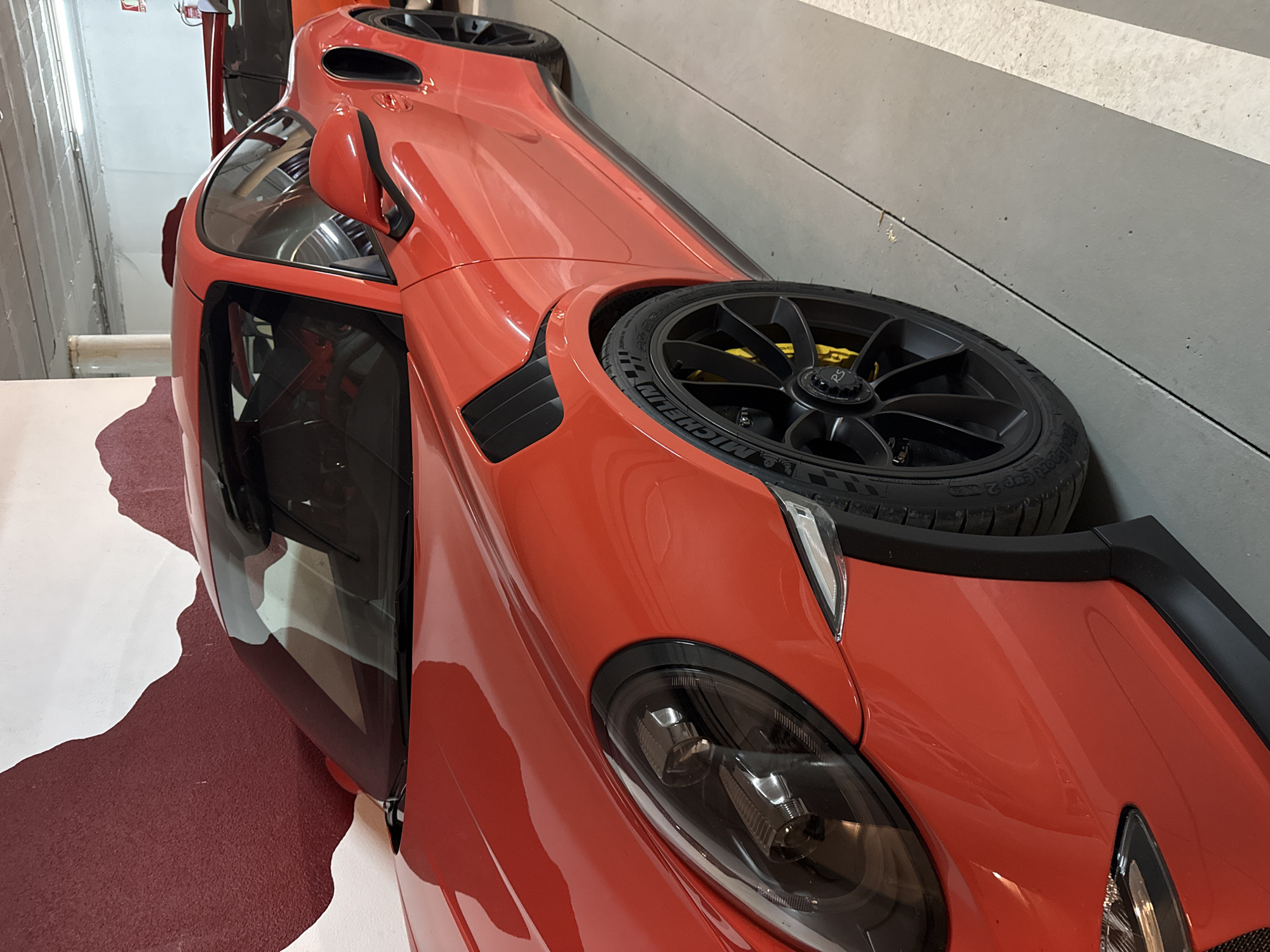A red sports car is photographed at an angle indoors. The car has a black wheel and tire on its hood near the passenger side, and the front headlight is visible. The car is parked on a floor that has a white and burgundy pattern. The wall behind the car is a light grey, and there are horizontal lines present in the wall. The location is not identifiable as Benasque, Spain based on visual elements. No people or interactions are depicted.