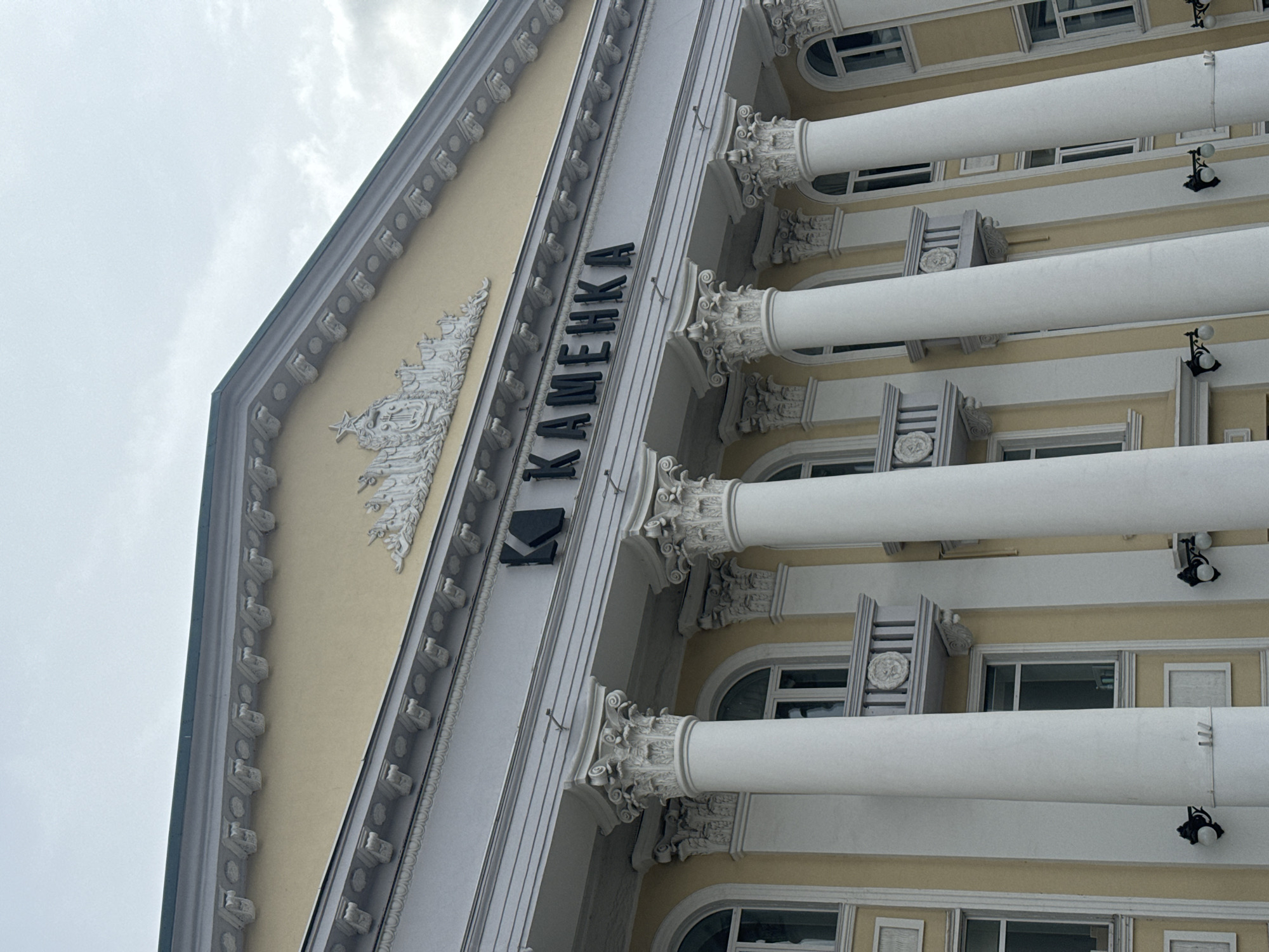 The image shows an exterior view of the "Kamenka" building in Krasnoyarsk, Russia. The architecture features classical elements, including columns with decorative capitals, a pediment, and ornamental detailing along the roofline. The facade is painted in light yellow and white tones. The name "KAMENKA" is displayed vertically in black letters on a white section of the building. The sky is overcast.