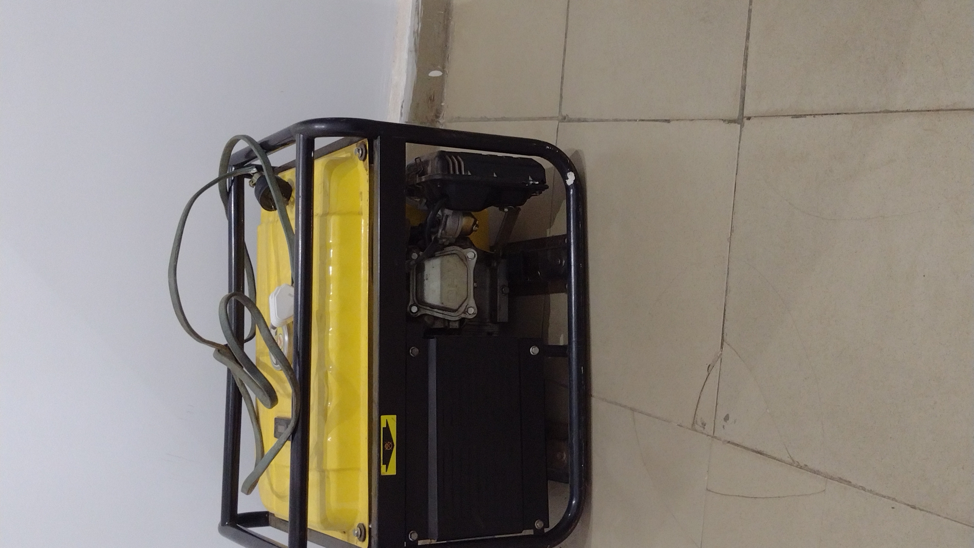 A yellow and black generator is situated on a tiled floor against a white wall. The generator has a black frame surrounding it and a yellow fuel tank. A gray electrical cord is wrapped around the frame on the left side. The tiles on the floor are light beige, and some have visible cracks. The wall is plain white. The location is Unknown, Unknown.