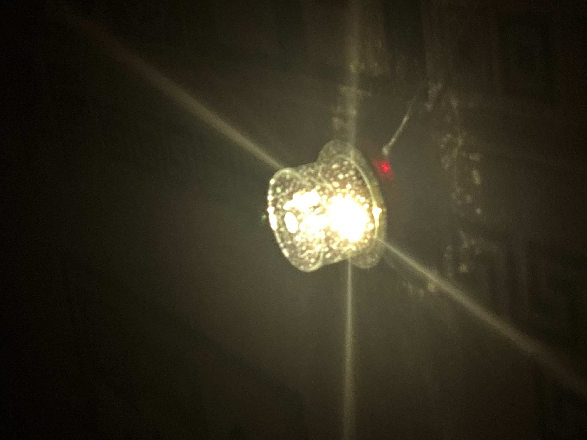 The image shows a close-up, low-light shot of a small, illuminated object that resembles a miniature top hat. The object is highly reflective and appears to be made of a transparent or translucent material with embedded reflective particles, giving it a sparkling appearance. A bright, warm light emanates from within the object, creating a strong glow. The object is situated on a dark surface, and light flares or beams are visible, extending from the object across the frame. There is also a small red light visible near the base of the object. The background is dark and indistinct, suggesting a nighttime or dimly lit environment. The scene appears to be an indoor setting, possibly a room, given the reflections and lighting. There is no visible indication of the specific location within Kryvyi Rih, Ukraine.