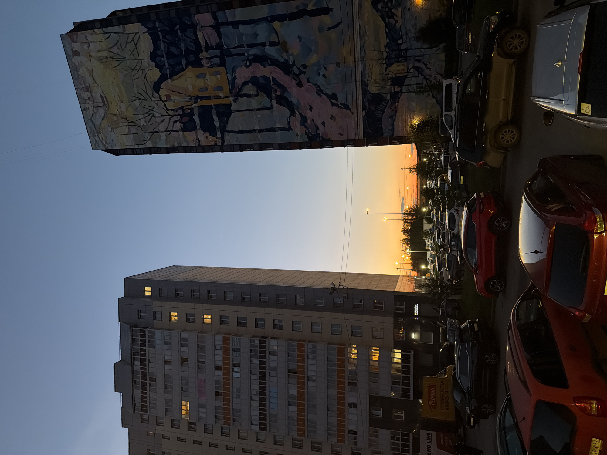 The image shows an exterior view of two apartment buildings and a parking lot filled with cars at dusk. The sky is a gradient of blue and orange, indicating sunset. One building on the left is a modern, multi-story apartment complex with many windows, some of which are illuminated from within. The other building on the right is taller and features a large mural on its side, depicting a natural scene with trees and a house. In the foreground and midground, numerous cars are parked in an organized lot. Streetlights and building lights are visible, contributing to the illumination of the scene. The environment appears to be a residential urban area. There are no people visible in the frame.