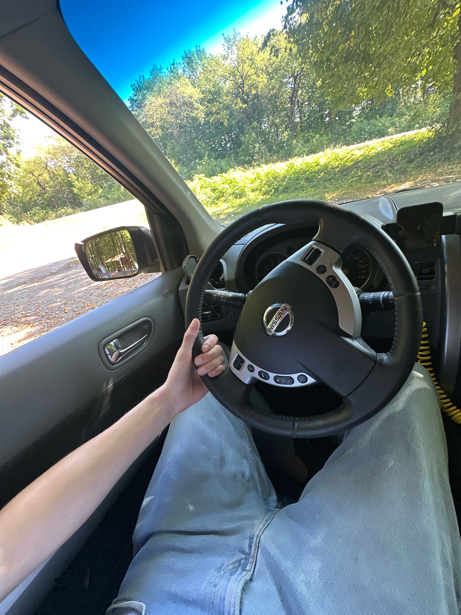 The image shows the interior of a car, with a person's hand on the steering wheel. The steering wheel is black leather with silver accents and the Nissan logo in the center. The dashboard has a speedometer and other gauges visible. The driver is wearing light blue jeans. The view through the windshield shows a tree-lined road and dappled sunlight. The side mirror reflects a structure that appears to be a picnic shelter. The scene suggests a drive through a rural or park-like area. There is no specific indication of the city of Shatava, Ukraine.
