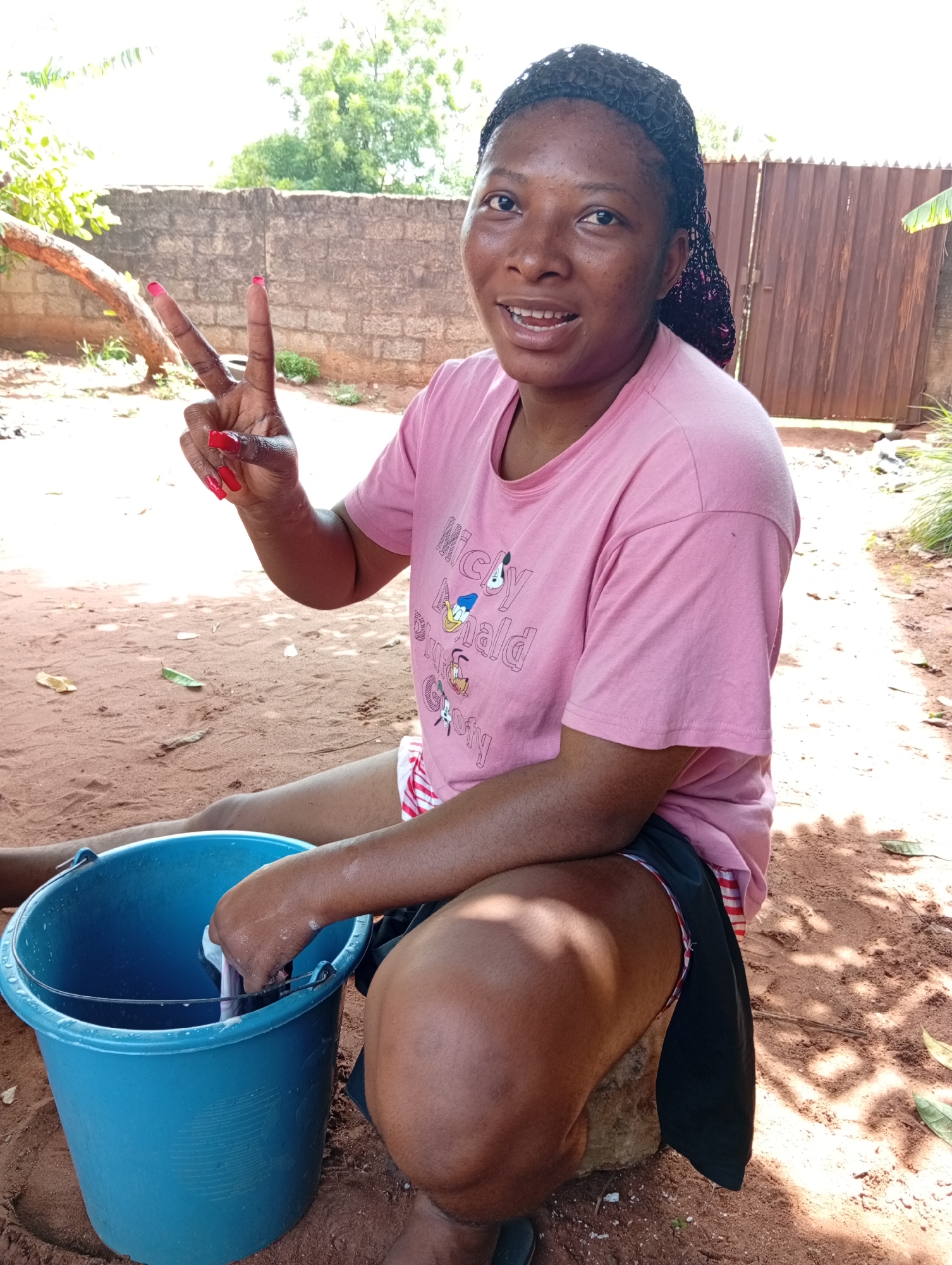 A young adult woman is pictured outdoors in what appears to be Ogidi, Nigeria, sitting or squatting on reddish-brown earth. She has dark skin and her dark hair is styled in braids. She is wearing a light pink t-shirt adorned with cartoon characters, specifically "Mickey," "Donald," and "Goofy" visible on the front. Underneath, she wears red-and-white striped shorts, with a black cloth draped over her lap.
Her left hand, adorned with bright red nail polish, is raised in a peace sign towards the camera. Her right hand is immersed in a large, light blue plastic bucket, which contains water and what looks like a fish she is handling. She is smiling broadly and looking directly at the viewer, conveying a cheerful and friendly expression.
The background features a low, weathered brick wall, behind which a brown corrugated metal gate is partially visible. Lush green trees and vegetation are present in the distance and to the sides, suggesting a natural or semi-rural setting. The bright lighting and visible shadows indicate it is daytime, likely sunny. The woman appears to be engaged in the activity of cleaning or preparing the fish in the bucket.
