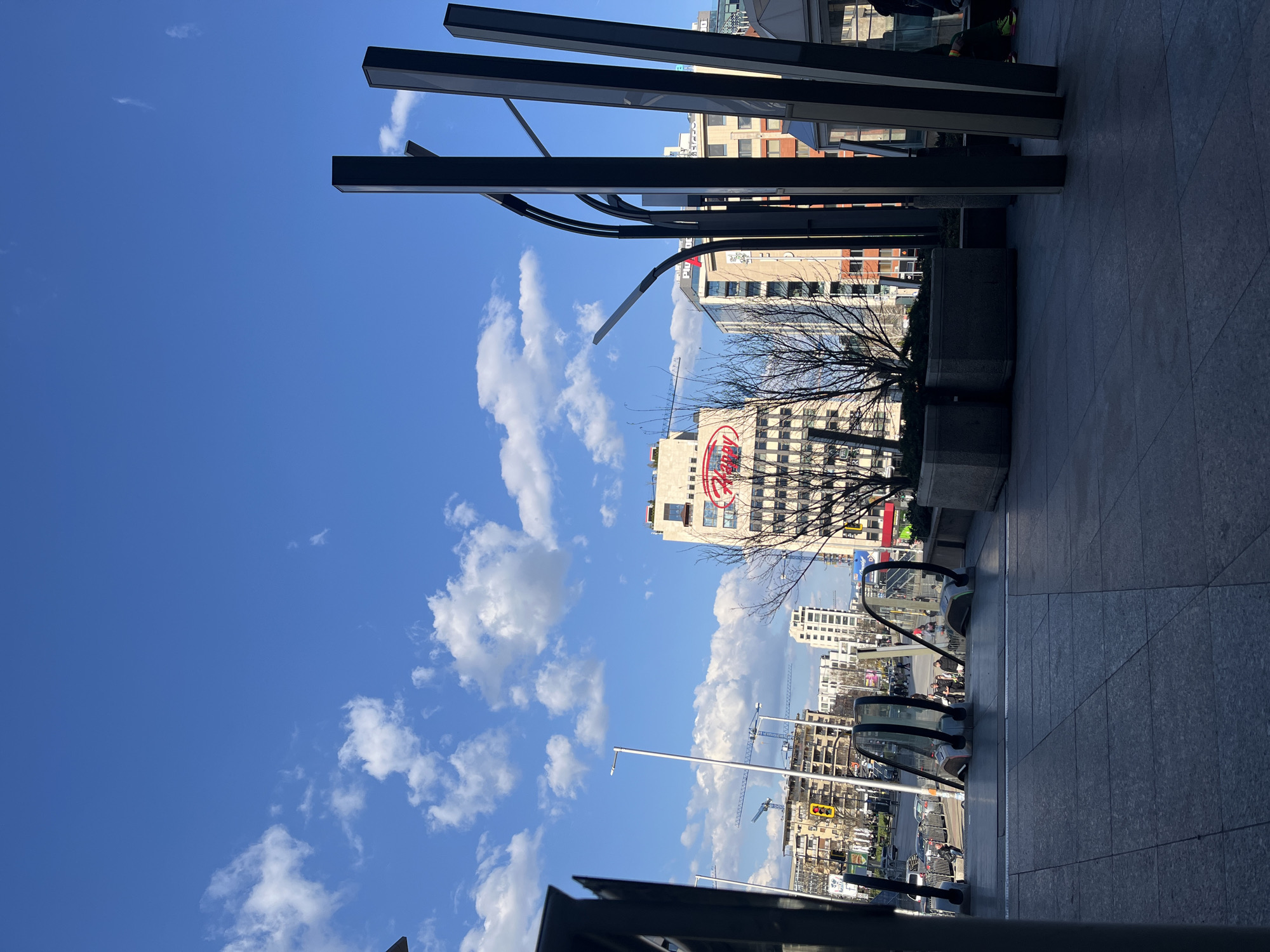 The image is an eye-level, outdoor shot in Sofia, Bulgaria, angled upwards towards the sky. The clear blue sky is dotted with fluffy white clouds. In the mid-ground, buildings of varying heights and architectural styles can be seen, including one prominently displaying a "Hilton" logo. Construction cranes are visible, suggesting ongoing development in the area. In the foreground, an escalator leads downwards, partially obscuring the view of the street. People are visible on and around the escalator. An array of metal structures and architectural elements frames the scene. The ground is paved with gray tiles.