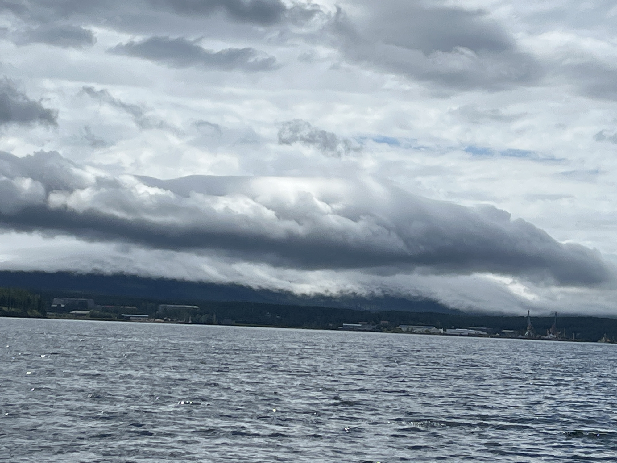 This image captures a wide view of a body of water, likely a river or bay, on a cloudy day. The water surface is textured with ripples and shows some scattered reflections. In the background, a dark, tree-covered shoreline is visible, stretching horizontally across the frame. Above the shoreline, a dramatic cloud formation hangs low, obscuring the sky and creating a moody atmosphere. The clouds are dense and varied in shade, with darker, heavier masses contrasting with lighter, wispy sections.
The location is identified as Zavety Il'icha, Russia. On the shoreline, there are hints of industrial or urban development, with what appear to be buildings and possibly cranes visible in the distance on the right side of the frame. The overall scene is natural and serene, dominated by the interplay of water and sky, with the distant shore providing a sense of place and human presence. There are no people or animals directly visible in the image. The lighting is diffused due to the overcast conditions, resulting in muted colors and soft shadows.