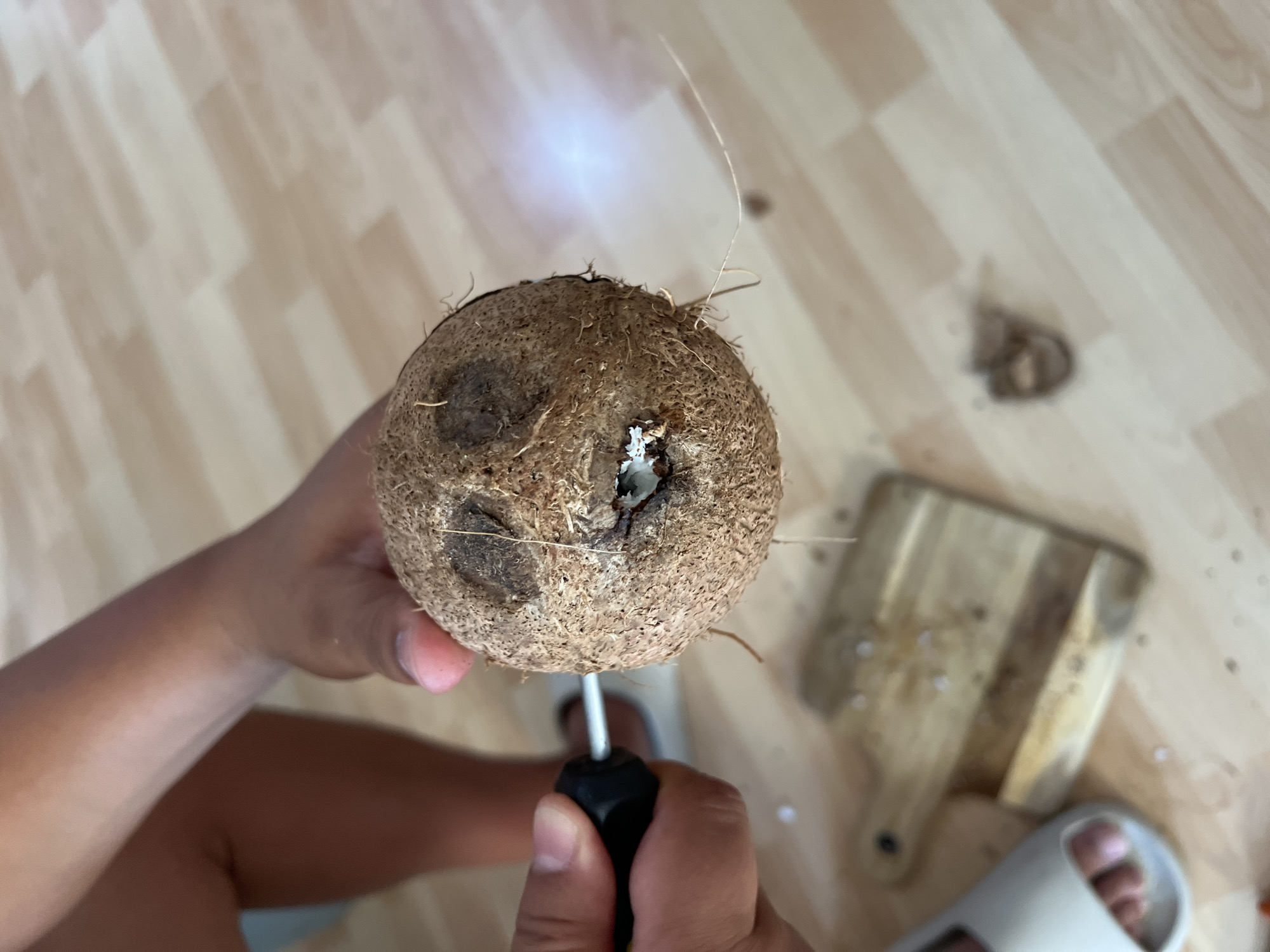 A person is holding a coconut and using a screwdriver to open it. The scene is indoors, on a light-colored wooden floor. A wooden cutting board with coconut pieces is visible in the background. The person is wearing light-colored sandals. I cannot provide the location because it is not provided in the image and is not possible for me to estimate.