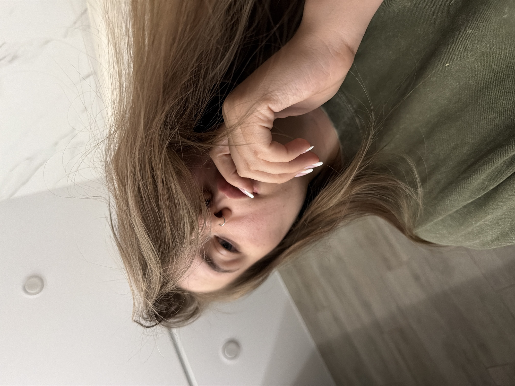 A vertically oriented close-up selfie of a young woman with light brown hair and fair skin. Her face is partially obscured by her hand with white-tipped nails. She has a nose ring on the left side of her nose. She is wearing an olive-green shirt. The background consists of a white wall with a light switch and light wood paneling. There is insufficient information to determine if the picture was taken in the city of Chaikovsky, Russia.