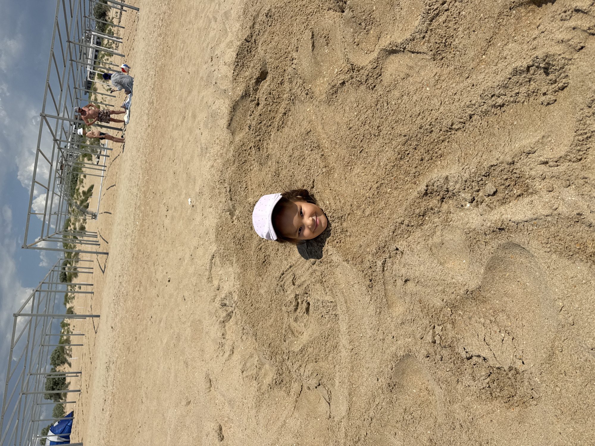 A young child, wearing a white baseball cap, is buried in sand up to their head on a beach. The child is smiling and looking towards the camera. The sand is light brown and textured with footprints. In the background, a metal structure, possibly a canopy frame, stretches along the beach. Two young men, one in a grey shirt and the other in patterned shorts, are visible near the structure, along with a beach ball. The scene suggests a sunny day at the beach in Vityazevo, Russia.