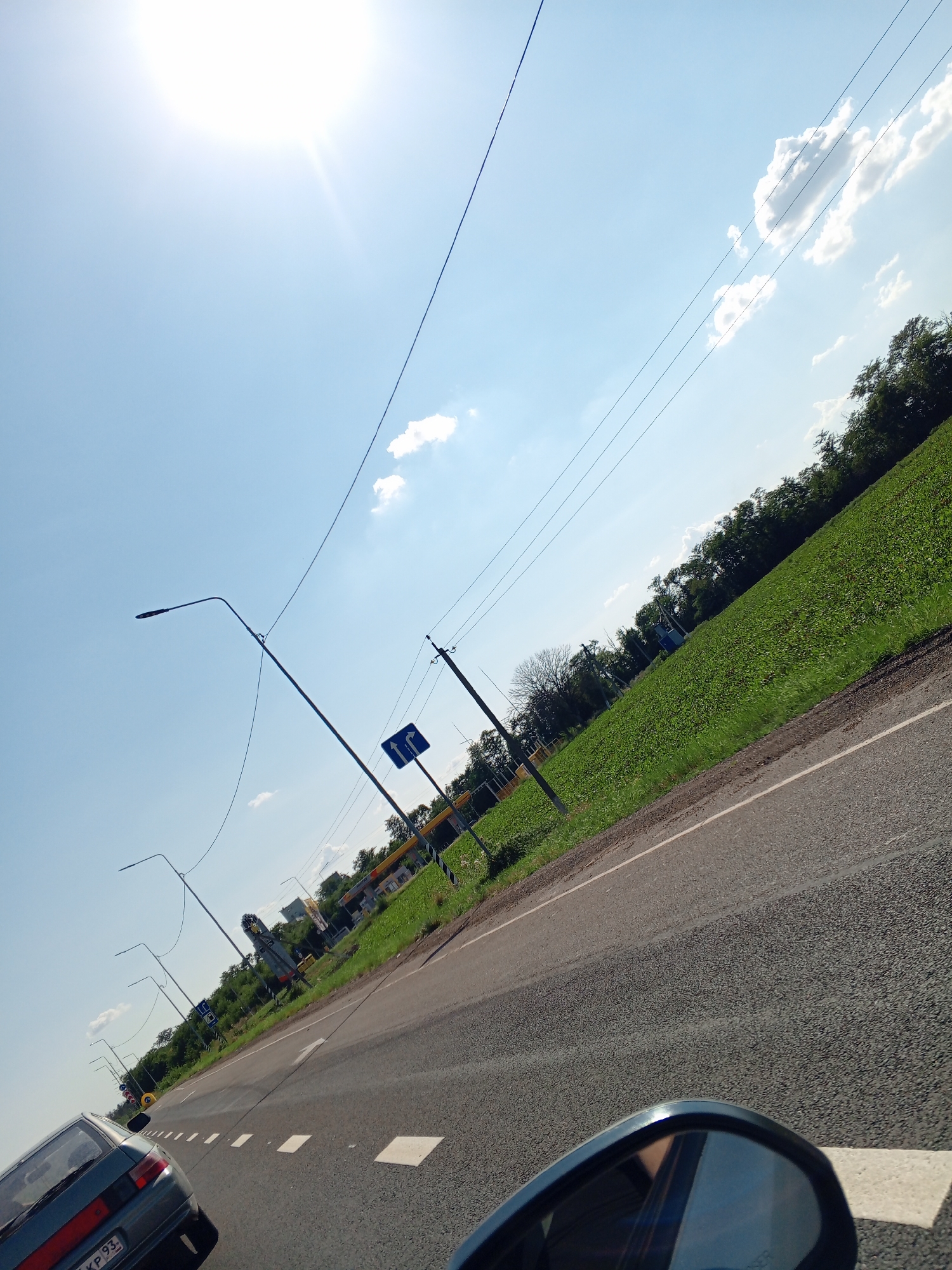 The image depicts a sunny day with a bright sun in the upper left corner and a clear blue sky with scattered clouds. A road stretches diagonally across the frame from the bottom to the upper right, with a green field bordering it on the right. A car is visible in the foreground, its side mirror reflecting the scene. Streetlights and power lines run parallel to the road. In the distance, there appears to be a gas station or a commercial building. A blue road sign with white arrows indicating traffic direction is also present. The overall impression is that of a roadside view on a clear day.