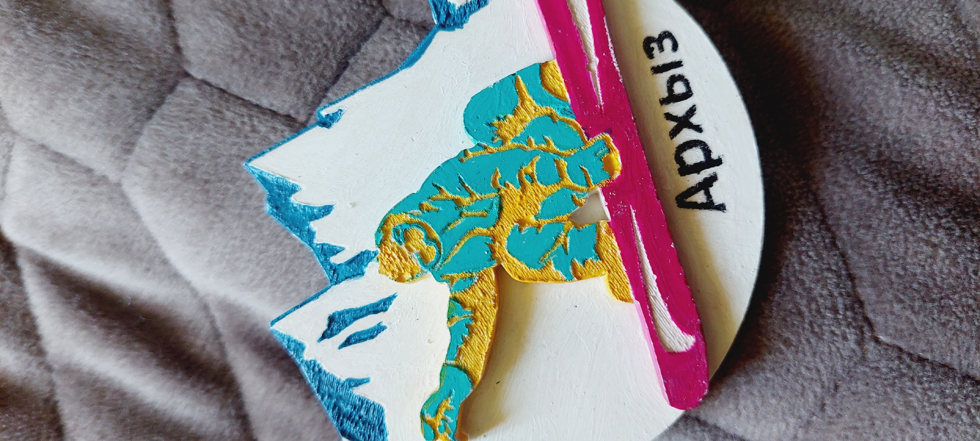This is a close-up, top-down shot of a circular, embossed magnet or ornament. The ornament is white and features a raised design that appears to be an abstract representation of a person, possibly a skier, in motion. The figure is depicted with a turquoise body outlined and accented with gold. Parts of the figure and surrounding elements are also colored white and a vibrant pink, suggesting movement or energy. The background of the ornament is a textured, gray surface, likely fabric like a blanket or couch.
The text "Архыз" is written in black, stylized Cyrillic letters along the right edge of the ornament, oriented vertically and readable from bottom to top. The inscription suggests a connection to Arkhyz, a resort town in the Karachay-Cherkess Republic of Russia, known for its mountain scenery and winter sports. The overall impression is that of a souvenir item, possibly commemorating a trip to this location. The lighting is neutral, with no strong shadows, and the focus is sharp on the ornament and its details.