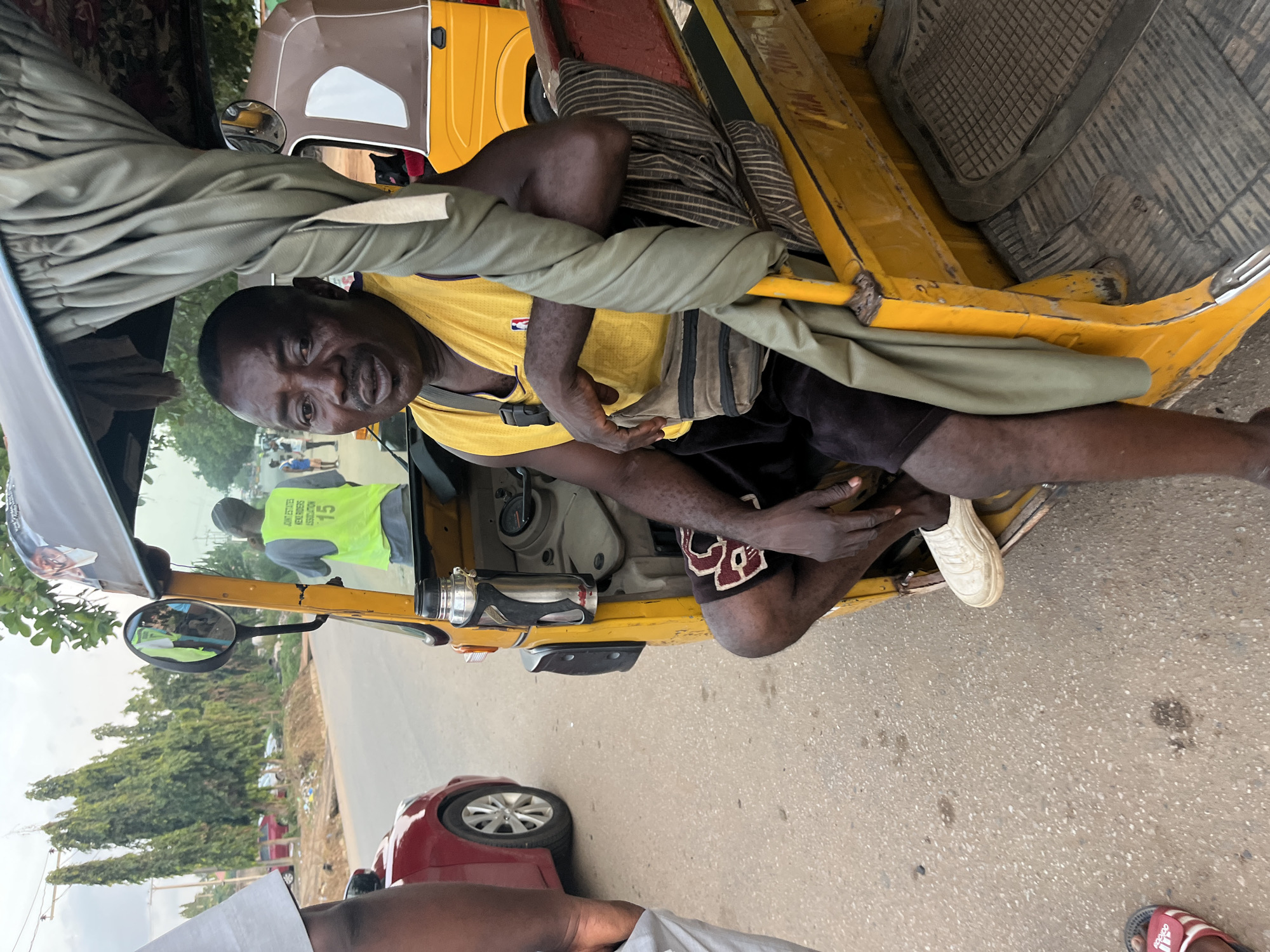 A man sits in the driver's seat of a yellow auto-rickshaw on a paved street, likely in Abuja, Nigeria. He wears a yellow sleeveless vest over a purple t-shirt, dark shorts, and white slip-on shoes. A grey bag with a black strap is slung across his chest. The auto-rickshaw has a tan-colored roof and side panels, and a grey fabric curtain hangs from the roof. In the background, other auto-rickshaws and a red car are visible, along with trees lining the street. A person wearing a reflective yellow vest with "JOINT ESTATES VENDORS ASSOCIATION 15" printed on the back is seen in the driver's side mirror of the auto-rickshaw. The weather appears overcast.