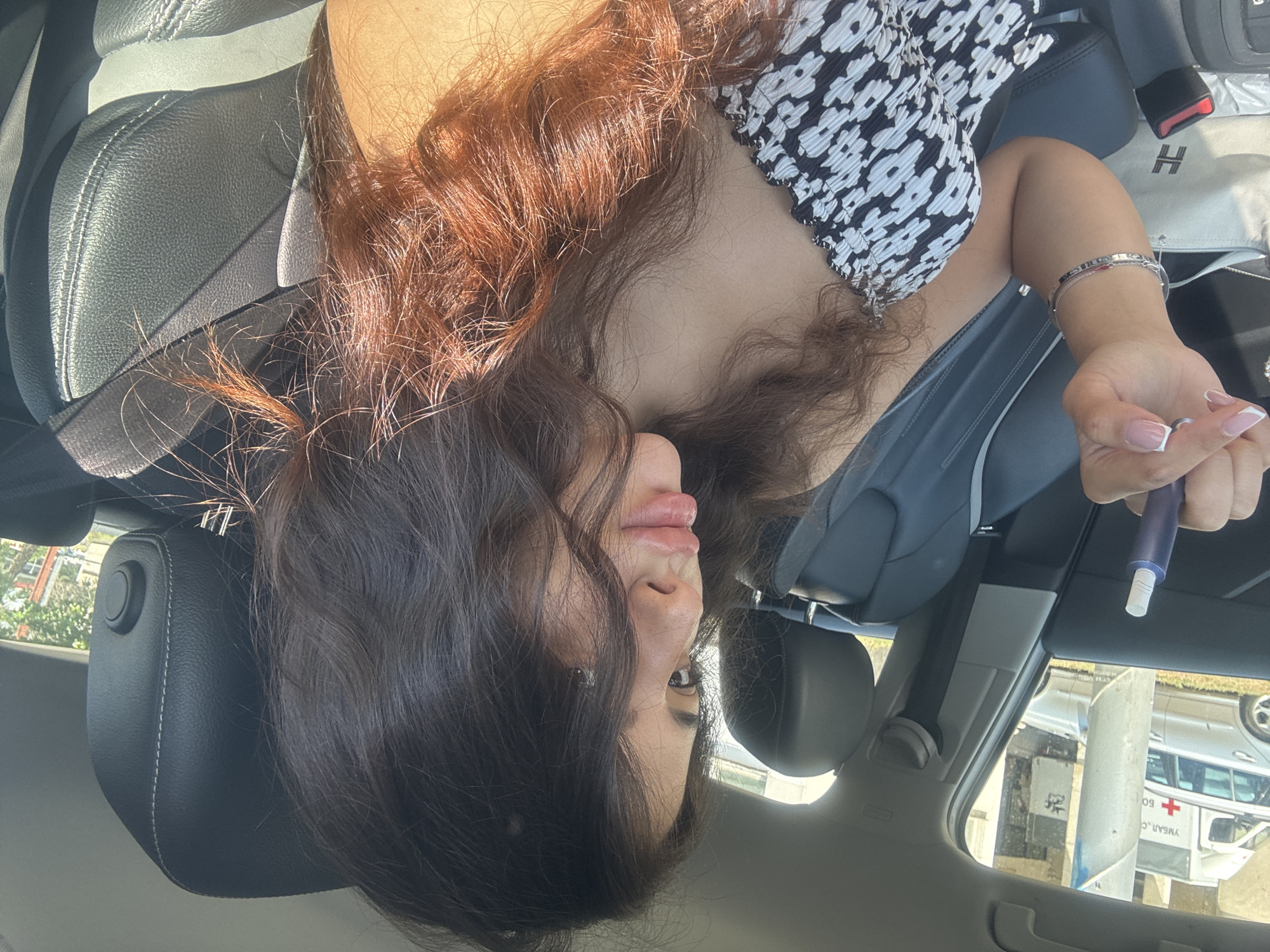 A young woman with dark, wavy hair is sitting in the passenger seat of a car, looking to her right. She is wearing a black and white patterned strapless top. Her right hand, which is adorned with French manicured nails and bracelets, is holding a blue and white object, possibly an e-cigarette. The car interior is visible, including a dark leather seat and seatbelt. Outside the car window, an ambulance with a red cross symbol and other vehicles are visible, suggesting the location might be near a hospital or medical facility in Plovdiv, Bulgaria. Sunlight is casting shadows on the car's interior and highlighting the woman's hair.