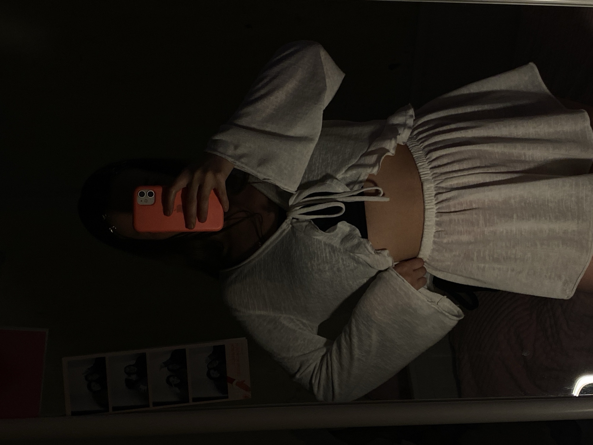 The image displays a mirror selfie, rotated 90 degrees counter-clockwise, taken in a dimly lit indoor setting, likely at night or in a dark room. The subject is a person, likely female, whose face is largely obscured by the coral-colored smartphone she holds in her right hand to take the picture. The smartphone features two distinct camera lenses on its back.
The person is wearing a light grey or white long-sleeved top with flared sleeves, ruffled details around the neckline, and ties at the front. This top appears to be a crop top or a shrug worn over a dark garment, revealing a portion of her midriff. She is also wearing a light grey or white skirt or bottom with a prominent ruffled, elasticated waistband.
In the bottom left corner of the reflection, a photo booth strip containing several small black and white photographs is visible, positioned next to a red rectangular object, possibly a book or a frame. The overall atmosphere is intimate and casual due to the low lighting and personal nature of a selfie. No visible text is discernible in the image, and there are no specific location cues to indicate the stated location of Votkinsk, Russia.