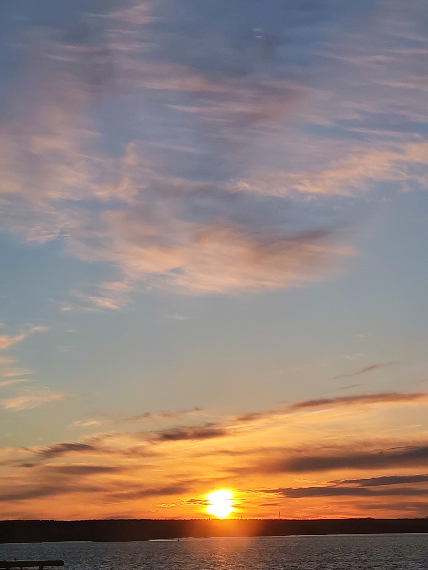 The image shows a sunset over water, likely a lake or river. The sky is filled with clouds, colored in shades of orange, pink, and blue by the setting sun. The sun itself is visible near the horizon, appearing as a bright spot. A dark treeline is visible on the horizon, separating the sky from the water. The water reflects the colors of the sky. The location is likely near Bol'shie Mkhi, Russia, based on the prompt.