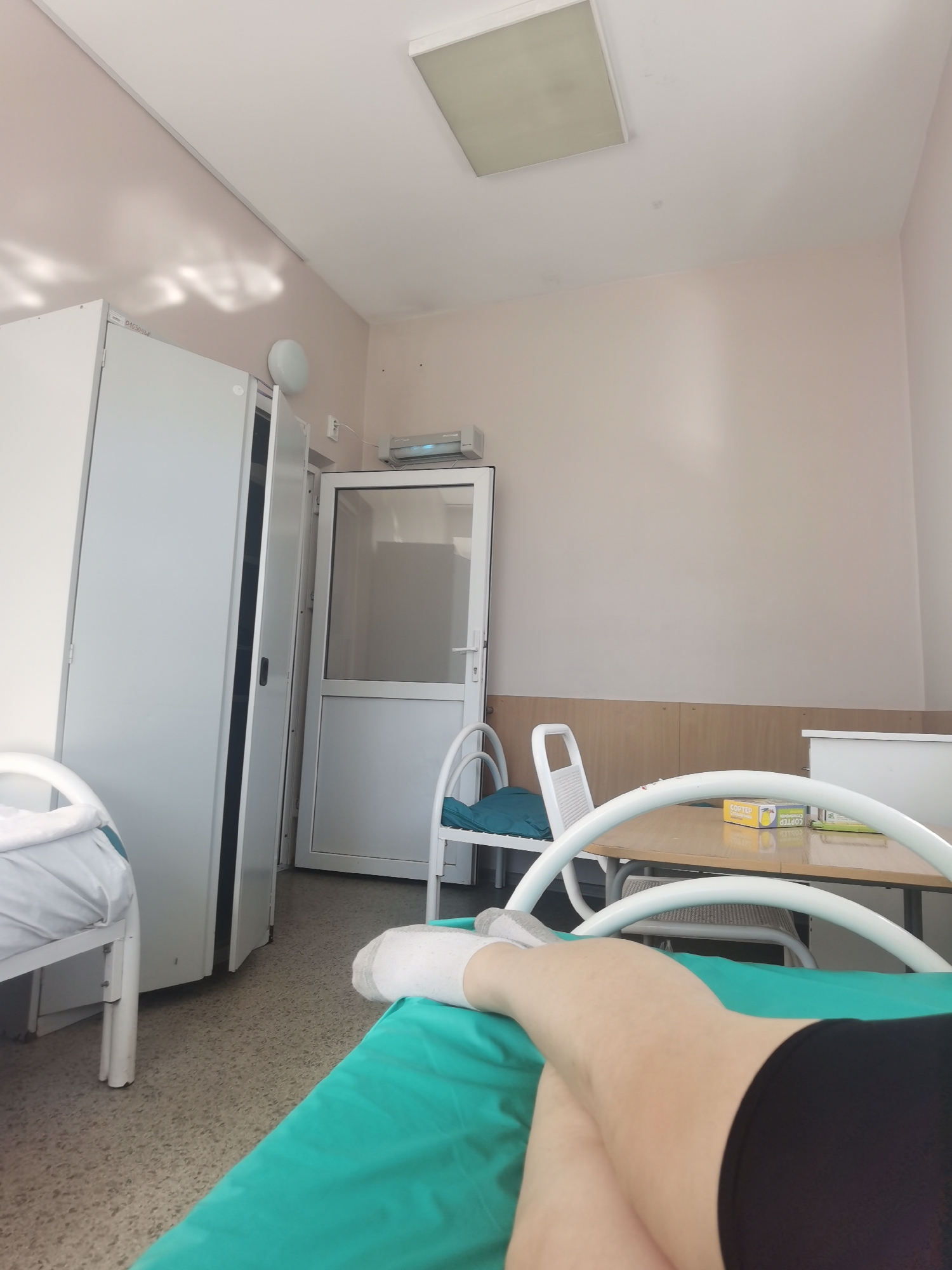 The image depicts the interior of what appears to be a hospital room in Perm, Russia. A person, likely a patient, is lying on a hospital bed with their legs crossed, wearing a white sock and black shorts. The bed has a green sheet. Adjacent to the bed is a small table with two boxes on top, labeled "COPTEP" in Cyrillic script. A white chair with a blue cushion is near the table. The room contains a white wardrobe, a white door, and a ceiling light fixture. The walls are painted light beige. The floor is covered with tiles. The exact location in Perm is not evident from the given image.