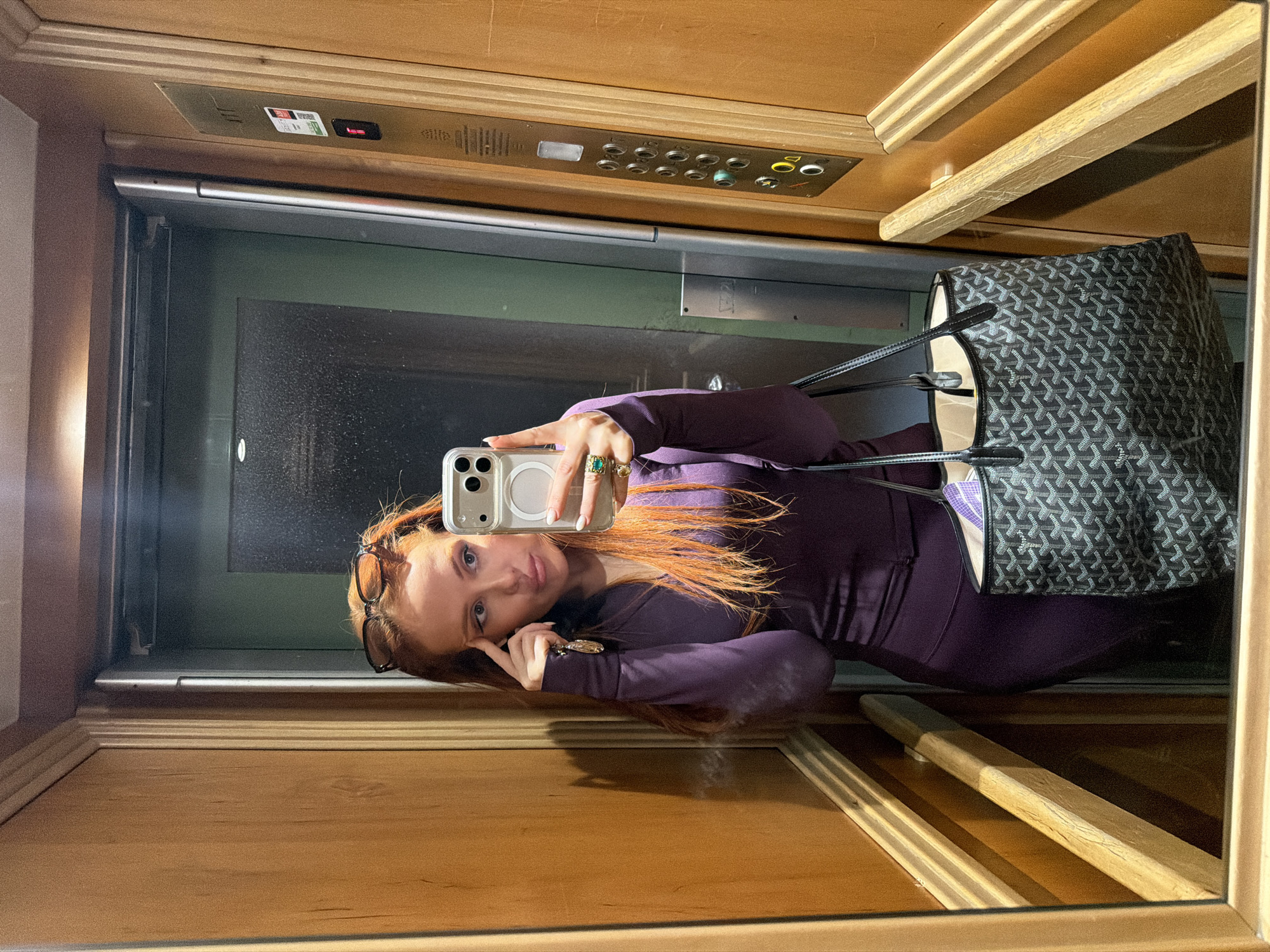 A woman is taking a selfie in an elevator. She is wearing a long-sleeved purple top and eyeglasses are resting on her forehead. She has red hair and is holding her phone in her right hand. Her left hand is near her face. A black and grey tote bag with black handles is next to her. The elevator has wooden paneling and a mirrored wall. Elevator buttons are visible at the top of the frame. The city of Paris, France cannot be ascertained from the information provided.