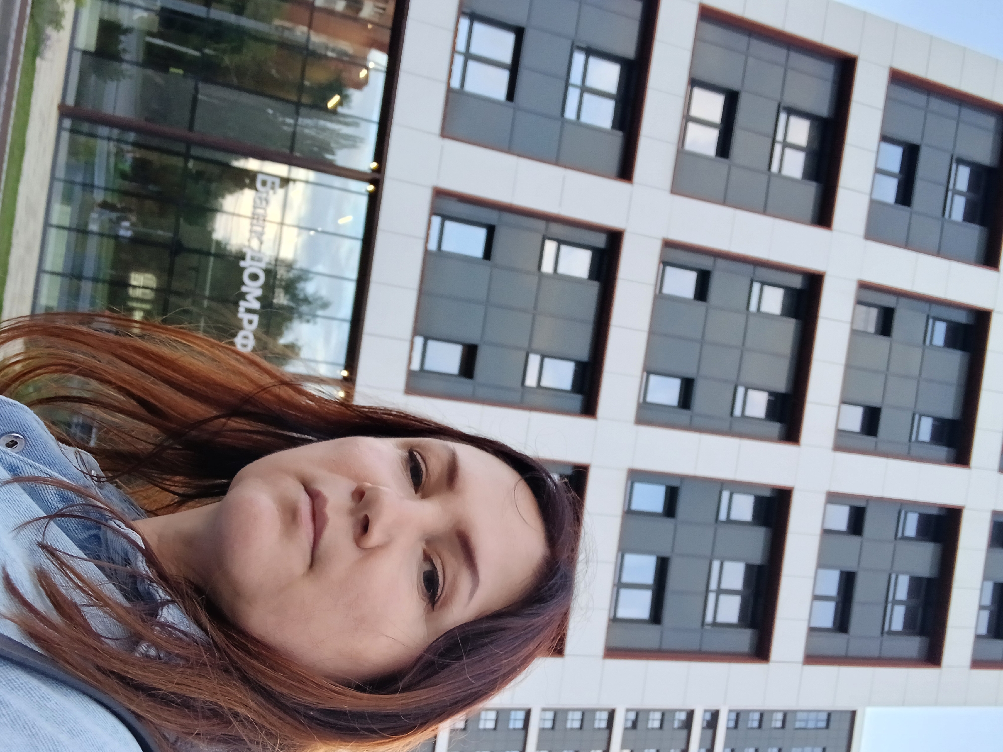 A woman with long, auburn hair and fair skin is captured in a selfie from the chest up. She is wearing a light blue denim jacket. Behind her, a modern building with a white facade and dark-paneled windows rises into a pale sky. The building's architecture features a grid of windows, with some of the panes reflecting the surroundings. In the lower part of the image, a glass-fronted entrance to a building is visible, displaying the white text "Бизнес ДОМ.РФ" (Business DOM.RF). The environment appears to be an urban setting, likely a business district or residential complex, given the contemporary architecture.