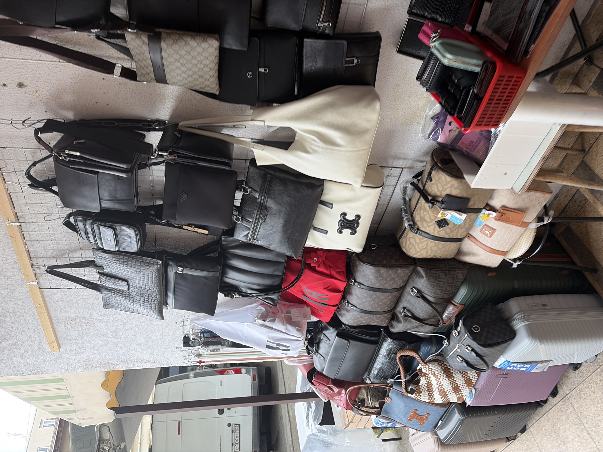 The image depicts an outdoor stall selling a variety of bags and luggage. The stall is located in Sochi, Russia, evidenced by the Russian license plate on a white van visible in the background. The stall displays an assortment of handbags, tote bags, and duffel bags in various colors, materials, and brands, including items with patterns resembling Louis Vuitton and Fendi. There are also several pieces of luggage, including rolling suitcases in different sizes and colors. In the foreground, a red basket holds wallets and cardholders, and a white box contains more accessories. The stall appears to be set up against a textured wall, with several bags hanging from metal hooks. No people are visible in the image.
