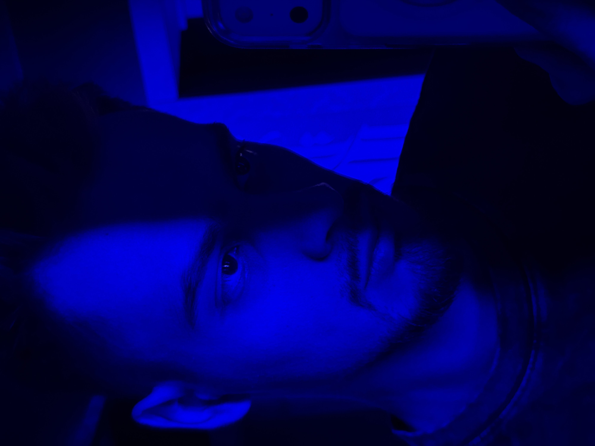 A selfie of a male with a light beard, taken in low light and dominated by a blue color cast. The man is holding a phone with multiple camera lenses visible, suggesting it is a modern smartphone. The location is impossible to discern based on the image. There is nothing present in the photo that suggests Krasnoyarsk, Russia.