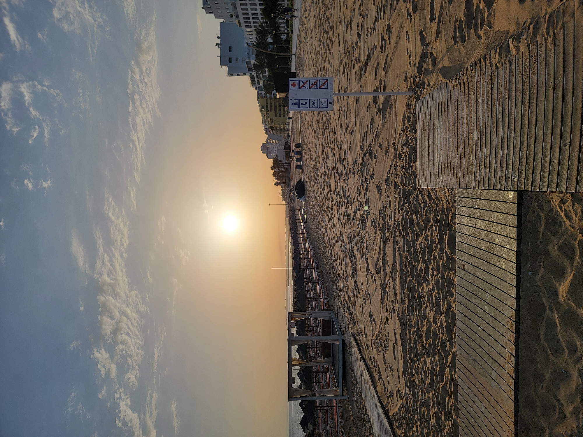 The image depicts a serene beach scene in Quarteira, Portugal, during what appears to be sunrise or sunset. The sun is low in the sky, casting a warm, golden glow across the sand and illuminating the scattered clouds. In the foreground, wooden walkways traverse the sand, providing access to the beach. To the left, a white, open-sided canopy structure with curtains stands on the sand. Beyond this, rows of umbrellas and lounge chairs are visible, suggesting a popular tourist destination. In the mid-ground, a signpost displays several pictograms, indicating rules or amenities available at the beach. Further in the background, the coastline is lined with multi-story buildings, characteristic of a coastal resort town. A few individuals are casually walking along the paved promenade beside the buildings. The overall atmosphere is tranquil, with the gentle waves of the ocean implied by the vast expanse of sand leading to the horizon.