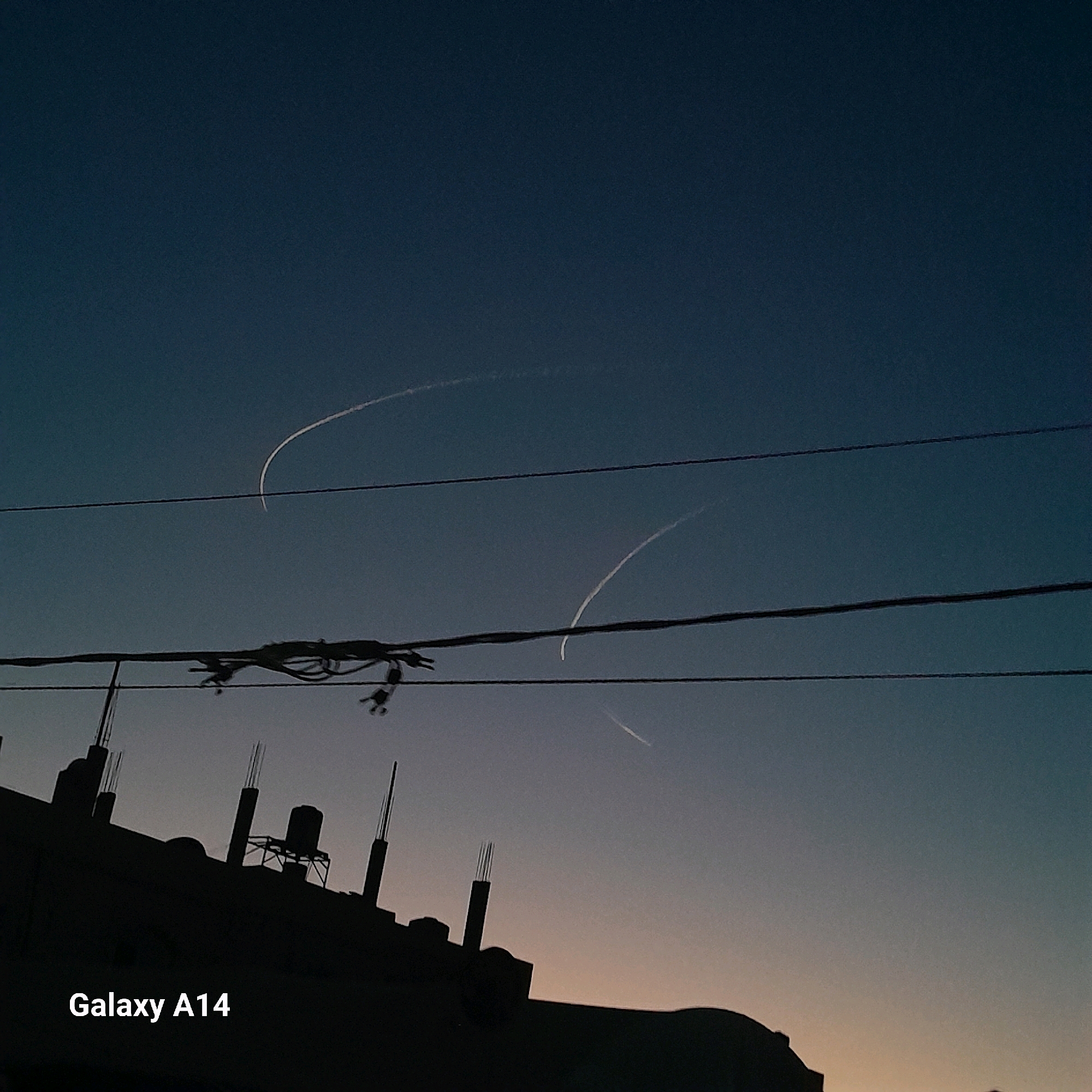 The image captures a twilight scene in Deir el-Balah, Palestinian Territories. In the sky, two faint contrails from aircraft arc across the deepening blue. Power lines stretch horizontally across the frame, one set lower and more prominent, with what appear to be birds perched on them. Below the sky and power lines, the silhouetted rooftop of a building is visible, featuring unfinished concrete structures and a water tank. The text "Galaxy A14" is overlaid in the bottom left corner of the image. The overall impression is one of a quiet, urban evening.