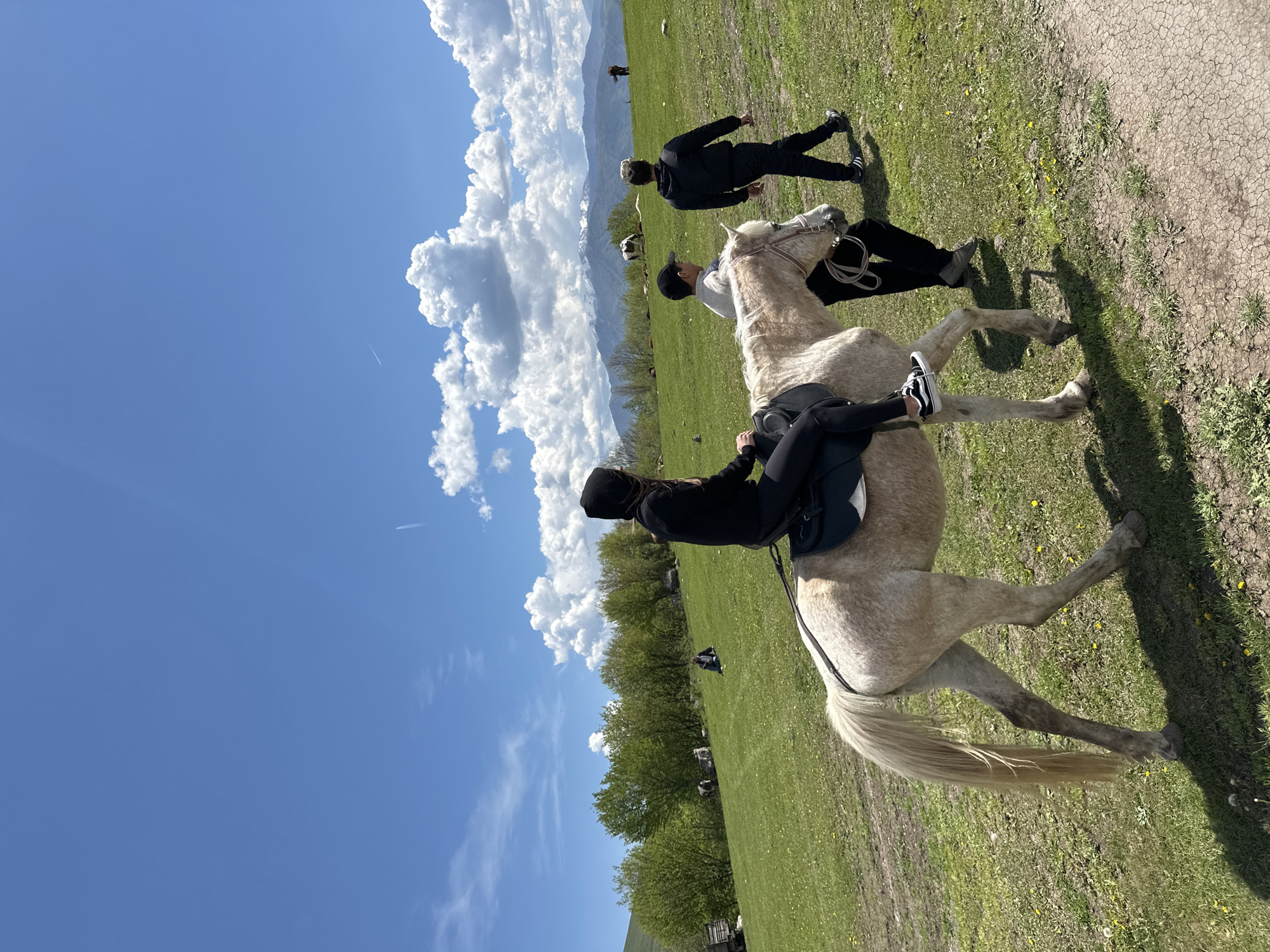 The image depicts an outdoor scene in Teghut, Armenia, featuring a pale-colored horse with a person riding it. The rider is wearing black clothing and a black hat. Another person in black clothing is standing nearby. There is a person walking in the background. The environment consists of a grassy field with trees and visible clouds in the blue sky. In the distant background, mountains can be observed.