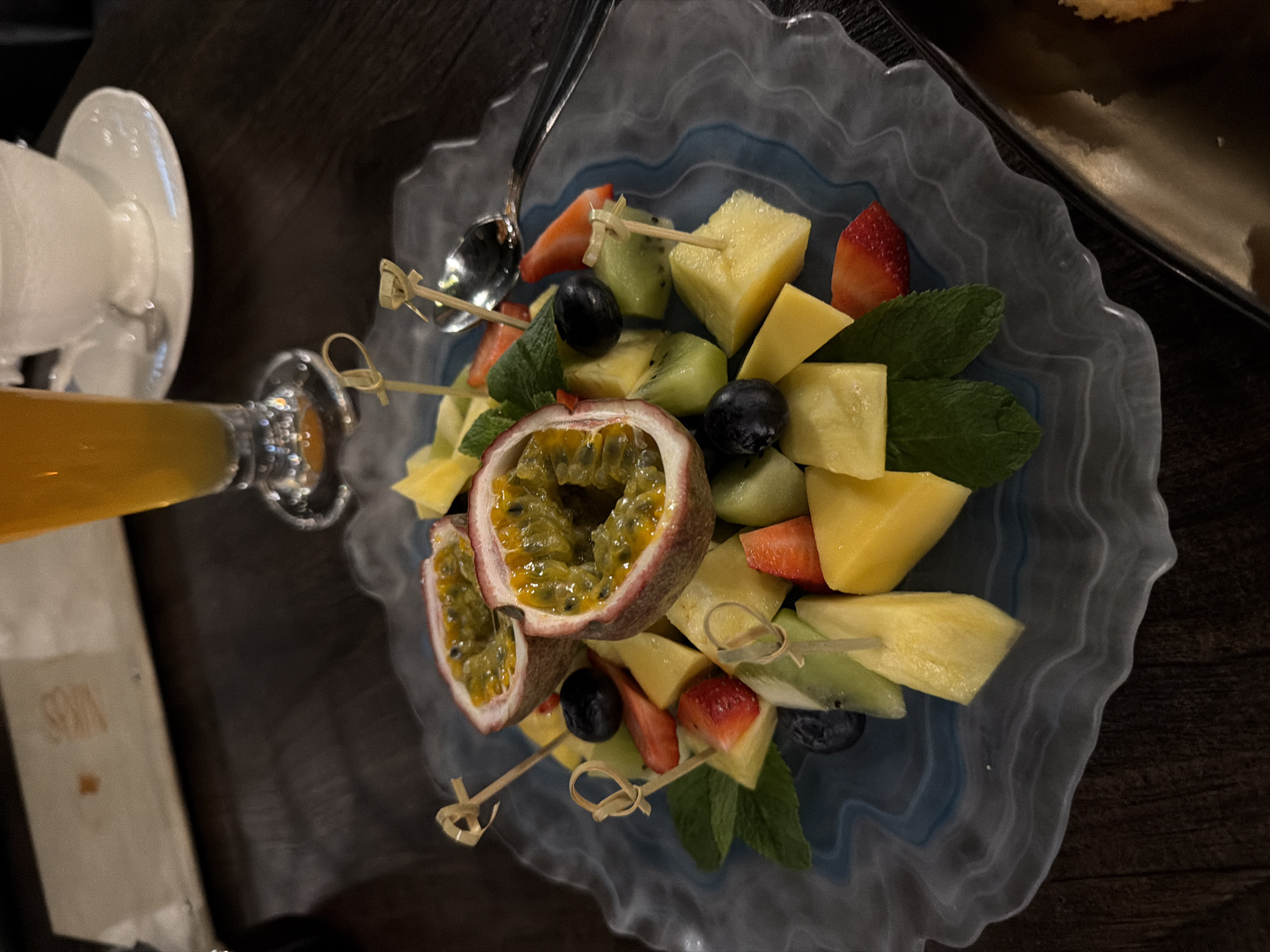 The image presents a close-up, top-down view of a fruit platter served in what appears to be an indoor dining setting. The fruit arrangement, served on a decorative, translucent blue-toned plate with wavy edges, features a mix of sliced mango, pineapple, kiwi, strawberries, blueberries, passion fruit cut in half, and garnishes of fresh mint leaves. Some of the fruit pieces are pierced with small wooden skewers. Adjacent to the plate is a glass of orange juice and a stack of white saucers. A small portion of another dish is visible in the upper right corner. The table surface is dark, possibly wooden, with a menu card or napkin visible near the glass. While the specific location cannot be determined, the menu card has "SIBYRS" printed on it, indicating that it is likely located in Siberia. It is, therefore, possible that the scene is from Krasnoyarsk, Russia.