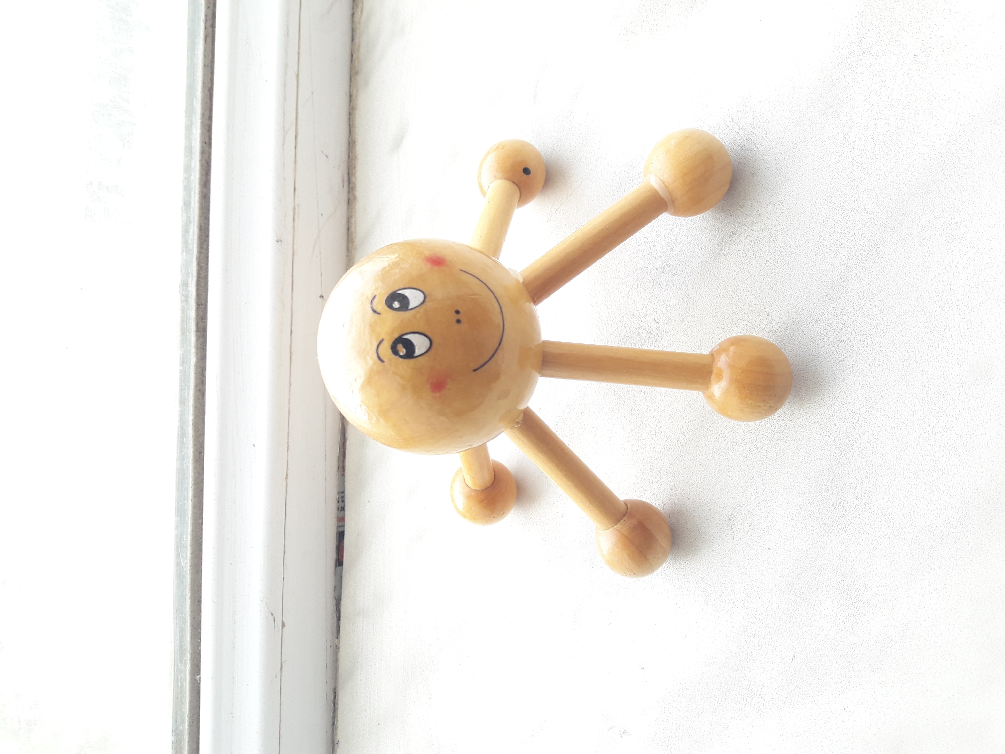 A wooden toy with a spherical body and four spherical feet sits on a white textured surface. The toy's body is painted with a smiling face, featuring two round eyes looking upwards, small dots for a nose, and a curved smile. Two small red circles are painted on its cheeks. The toy is positioned centrally in the frame, with its feet spread out. In the background, a white ledge or windowsill is visible, with a slightly darker line running across its surface, and beyond that, a bright, blurry white area which is likely an out-of-focus background or a window. The lighting is bright, suggesting it is daytime.