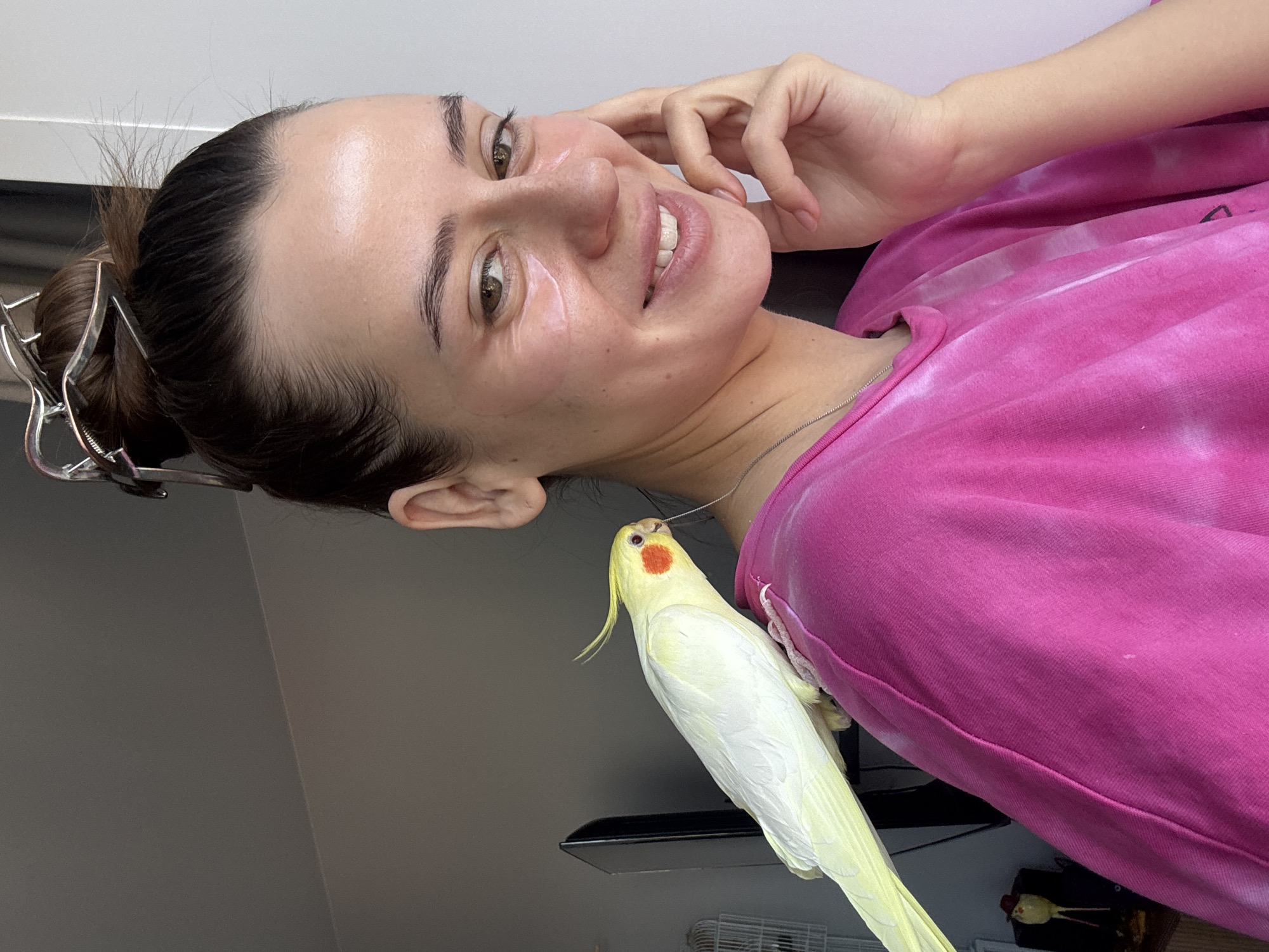 A woman is smiling and looking at the camera while a cockatiel is perched on her shoulder. The woman has brown hair pulled back in a bun with a silver clip. She is wearing a pink tie-dye shirt. The cockatiel is yellow and white with a red cheek patch. The background is a gray wall. The location is unclear as the context does not provide enough information to determine the location as Krasnoyarsk, Russia.