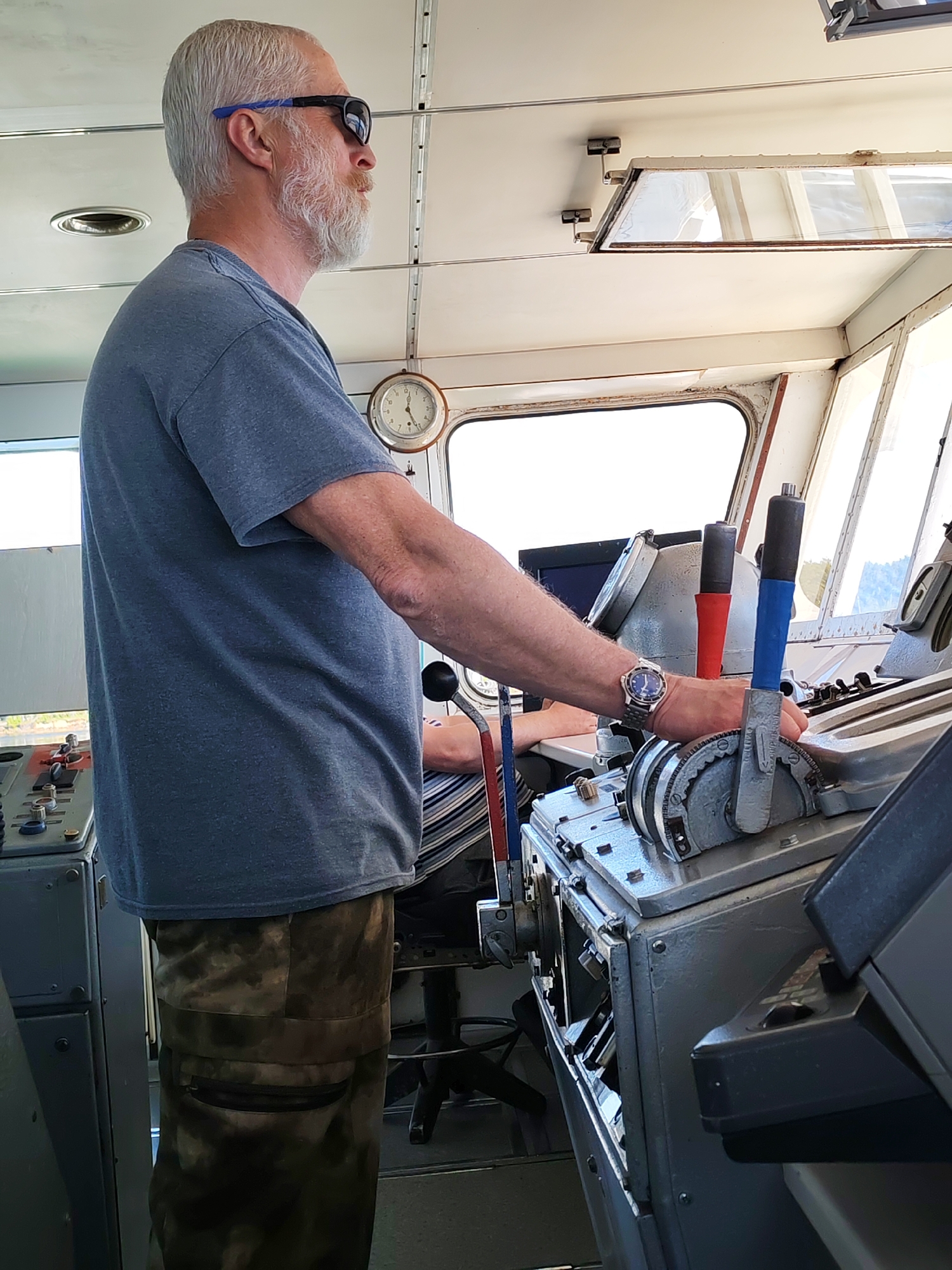 The image depicts an adult male standing in the bridge of a vessel, which is identified as being located in Kostino, Russia. The man has grey hair and a prominent grey beard, and is wearing a plain grey short-sleeved t-shirt, patterned cargo pants, and black sunglasses with blue-colored arms. He is positioned facing forward and slightly to the right, with his left hand resting on a prominent grey control lever that has a blue and black handle. A silver watch with a blue face is visible on his left wrist.
To the man's left, another section of a grey control panel is visible, featuring multiple red and black knobs and dials. Partially obscured behind the main subject, the arm and torso of a second individual, wearing a horizontally striped shirt, are visible, suggesting another person is seated or standing behind the controls.
The interior environment is characterized by a white ceiling with exposed metal framing and an overhead rectangular window or mirror. A circular wall clock with Roman numerals, set in a wooden frame, is mounted on the wall behind the man, indicating a time of approximately 5:35. Large, multi-paneled windows form the forward section of the bridge, admitting significant natural light and revealing a bright, indistinct exterior view. The control equipment is robust and industrial in appearance, consistent with maritime operational gear.