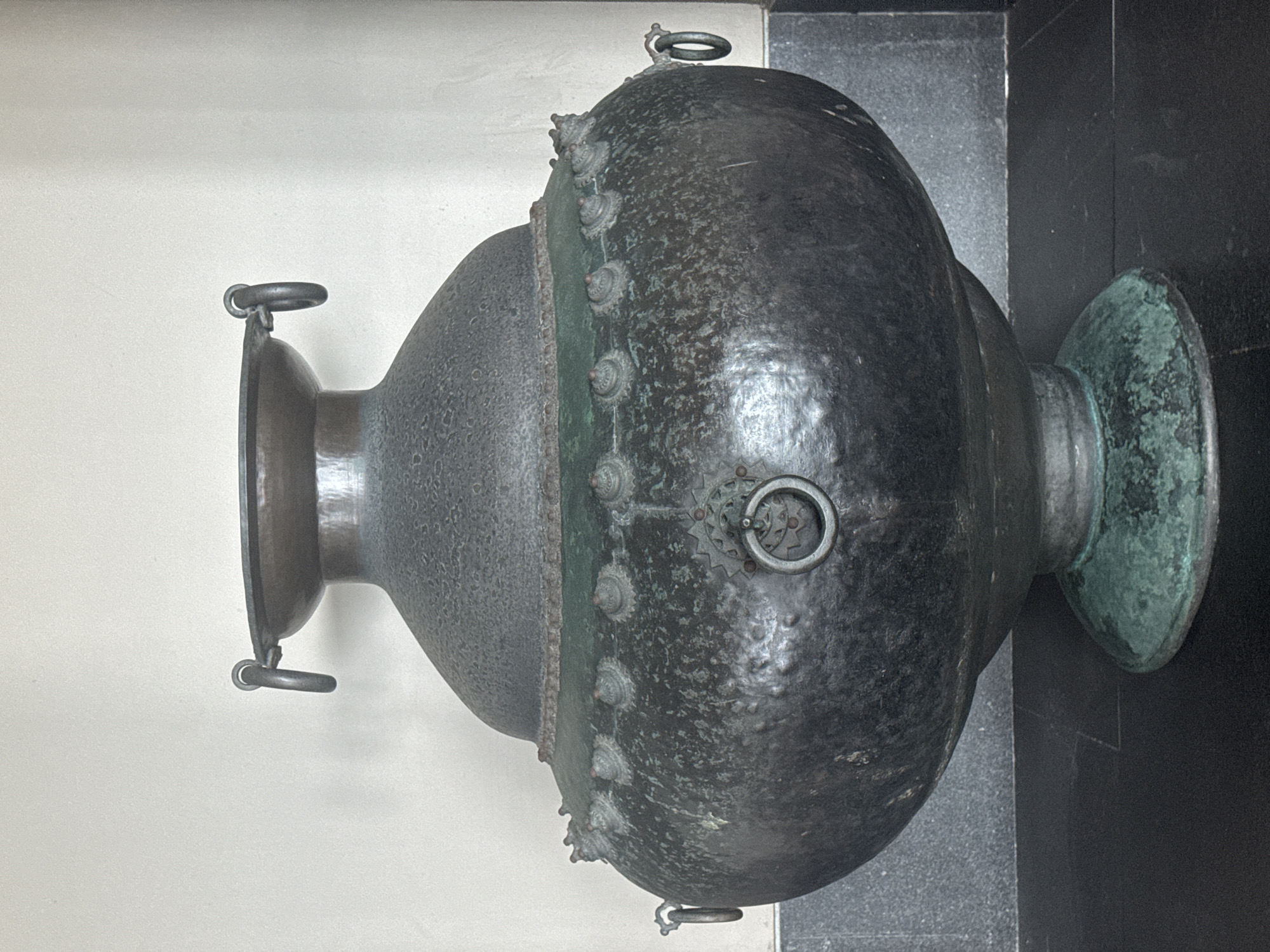 The image features a large, ornate metal vessel, likely an urn or cauldron, displayed upright in what appears to be an indoor setting. The object rests on a dark, polished surface, possibly stone or marble, against a plain, light-colored wall.
The vessel is composed of two main sections: a wider, bulbous lower body and a narrower, inverted bell-shaped upper part. A distinct horizontal band separates these sections, decorated with numerous conical studs or rivets, each featuring a textured circular base. The main body of the vessel has a dark, mottled texture, suggesting it is made of an aged metal like bronze or copper. A significant greenish patina is visible on the wide, flared base and parts of the connecting band, indicative of oxidation over time.
Several ring handles are integrated into the design. Two smaller rings are present near the rim of the upper section. The larger, lower section features a prominent circular ring handle on its side, backed by an intricate, rosette-like decorative plate. Another smaller ring is located near the bottom of this section. The overall appearance suggests a historical or antique artifact.
There is no activity taking place, as the object is a static display. No people are visible, and no text is discernible on the vessel or in the background. The scene is illuminated evenly, highlighting the object's details and textures. The location context for this media file is Kuta Selatan, Indonesia.