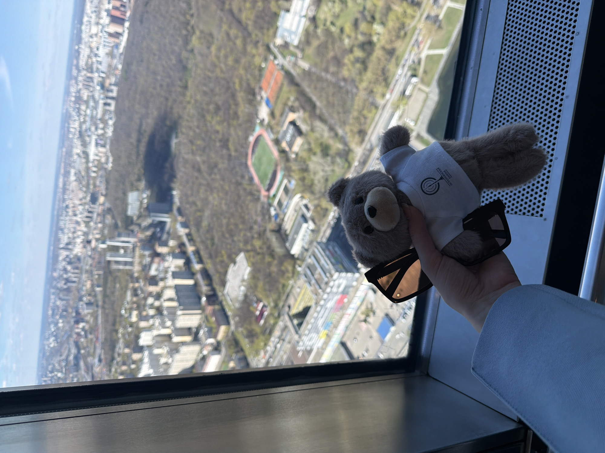 A person holds a small, gray teddy bear wearing a white shirt and sunglasses in front of a window, with an aerial view of Khimki, Russia, visible outside. The city stretches out to the horizon, with a mix of buildings and trees. A stadium with a red running track and green field is visible among the buildings. The window is set into a modern structure, with a perforated metal panel to the right. The person is wearing a light blue sleeve.