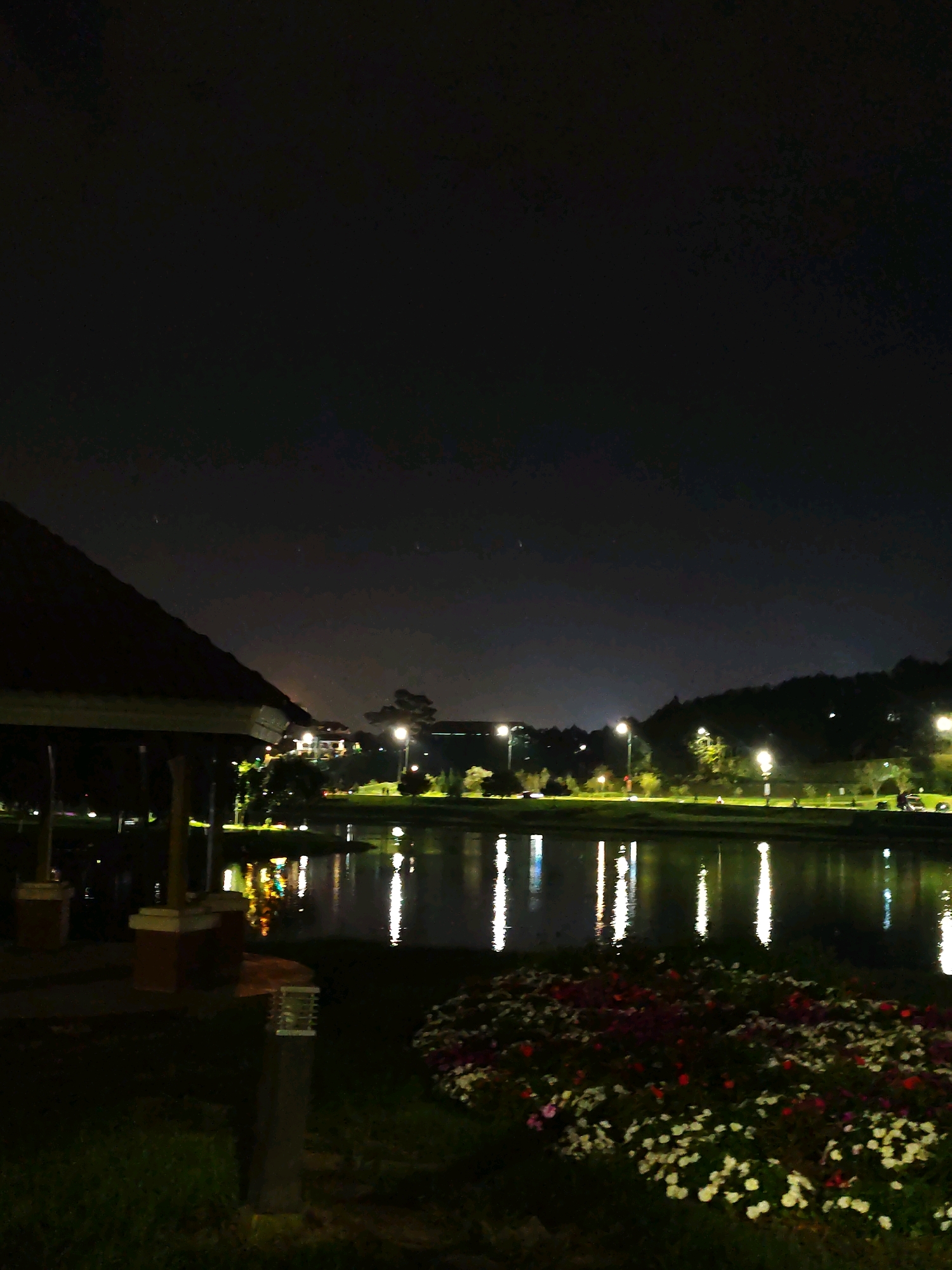 The image depicts a nighttime scene at Xuân Hương Lake in Đà Lạt, Vietnam. In the foreground, a covered pavilion with a dark roof is visible on the left, partially obscuring the view. A stone column supporting the pavilion is also present. To the right of the pavilion, a low, modern-style lamp post stands on a patch of grass. In front of this lamp post, a bed of flowers in various shades of red and white is illuminated.
The middle ground is dominated by the calm waters of Xuân Hương Lake, reflecting the lights from the opposite shore. The water surface shows streaks of light, indicating reflections from streetlights and buildings.
The background features a tree-lined embankment around the lake. Several lampposts with visible light sources line the path along the embankment, casting a warm glow. Buildings are visible further up the slope, also illuminated by lights. The distant hills are silhouetted against the dark night sky. The sky itself is dark with a few faint, distant stars visible. No people are clearly discernible in the scene.