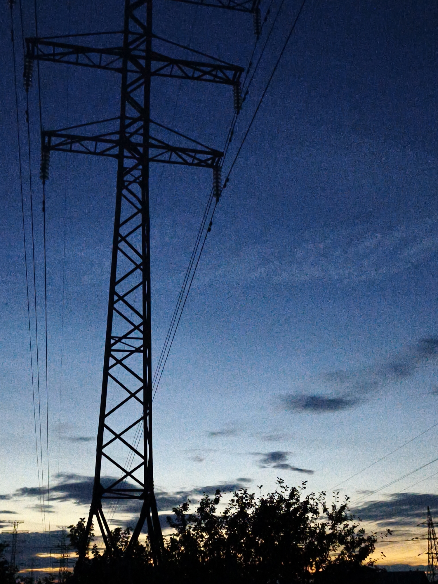 The image captures a silhouette of a tall, industrial electricity pylon against a darkening blue sky. The pylon is a prominent subject, characterized by its lattice structure and the power lines stretching from it. To the right of the main pylon, another, smaller pylon is visible in the distance, also silhouetted against the sky. In the foreground, a dark, dense silhouette of foliage from a tree or bushes partially obscures the base of the main pylon and extends across the bottom portion of the frame.
The sky displays a gradient of colors, transitioning from a deep, dusky blue at the top to lighter shades of blue and hints of orange and yellow near the horizon, suggesting either dawn or dusk. Scattered, wispy clouds are present in the sky, some appearing dark and textured, others lighter and more diffuse. The overall lighting indicates that it is either early morning or late evening.
No people are visible in the image. The setting appears to be outdoors, likely in a rural or semi-industrial area, given the presence of electricity pylons and open sky. The mood conveyed by the image is one of quiet and perhaps a touch of industrial starkness, emphasized by the silhouetted forms against the fading light.
There is no visible text within the image. The location context provided is Chaikovsky, Russia.
