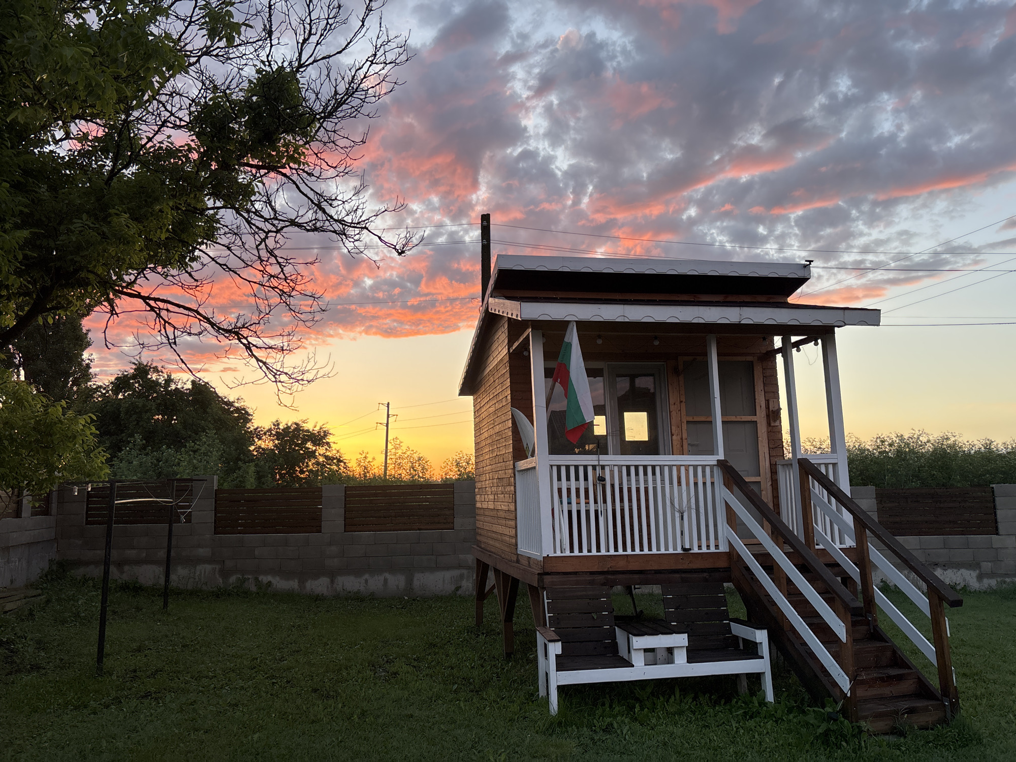 This image captures a tranquil evening scene at sunset in Pchelnik, Bulgaria. The sky is a dominant feature, displaying a spectacular blend of colors as the sun sets. The horizon glows with warm golden and orange hues, transitioning upwards into vibrant pink and red clouds that are scattered across the sky, eventually fading into lighter grey and soft blue tones.
The main subject of the photograph is a small, charming wooden house built on stilts, located slightly to the right of the center. It features a welcoming porch or deck with crisp white railings, and a set of sturdy wooden stairs with white and dark wood railings leads up to the entrance. Beneath the porch, a small, dark wooden bench with white accents offers a place to rest. A Bulgarian flag, identifiable by its horizontal stripes of green, white, and red, hangs vertically from the porch, providing a clear geographical cue. The house itself is constructed from horizontal wooden planks, lending it a rustic aesthetic, and a dark chimney pipe rises from its roof.
The foreground consists of a grassy yard, cast in the soft shadows of twilight. To the left, a simple metal structure resembling a clothesline stands in the grass. A sturdy fence forms the boundary of the property, built with grey concrete blocks at its base and topped with dark wooden horizontal slats. Beyond the fence, a line of dense trees and foliage is silhouetted against the bright sunset, with utility poles and wires visible in the distance. The scene is devoid of people or explicit activity, conveying a sense of serene peacefulness at dusk. No visible text is present in the image.