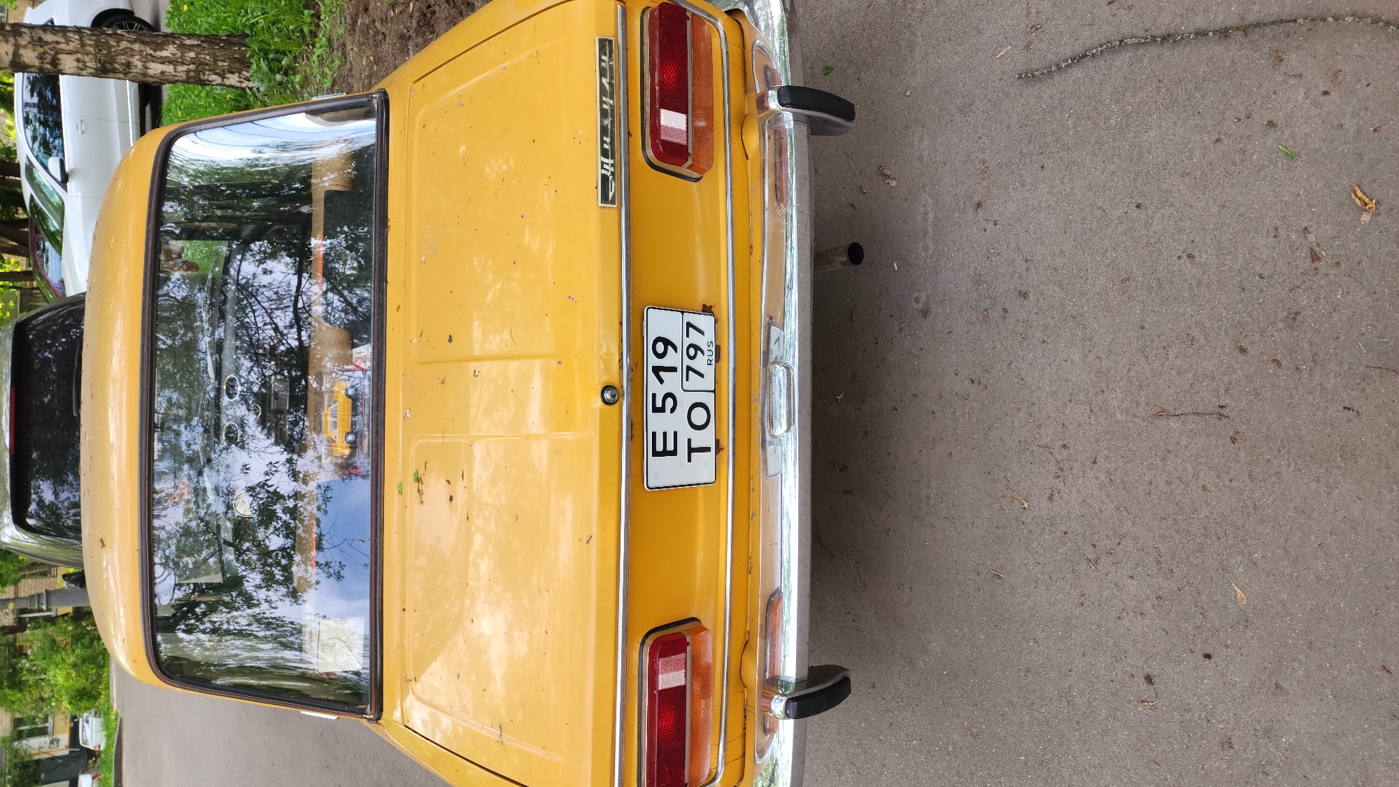 The image shows the rear of a yellow vintage car parked on a gray concrete surface. The car has chrome bumpers and a license plate that reads "E 519 TO 797 RUS." The car is parked next to a curb with some green foliage visible in the background, suggesting an urban setting, likely in Moscow, Russia, given the "RUS" on the license plate. Reflections of trees and buildings are visible in the car's rear window. The image suggests a quiet street or parking area, with no people directly visible.