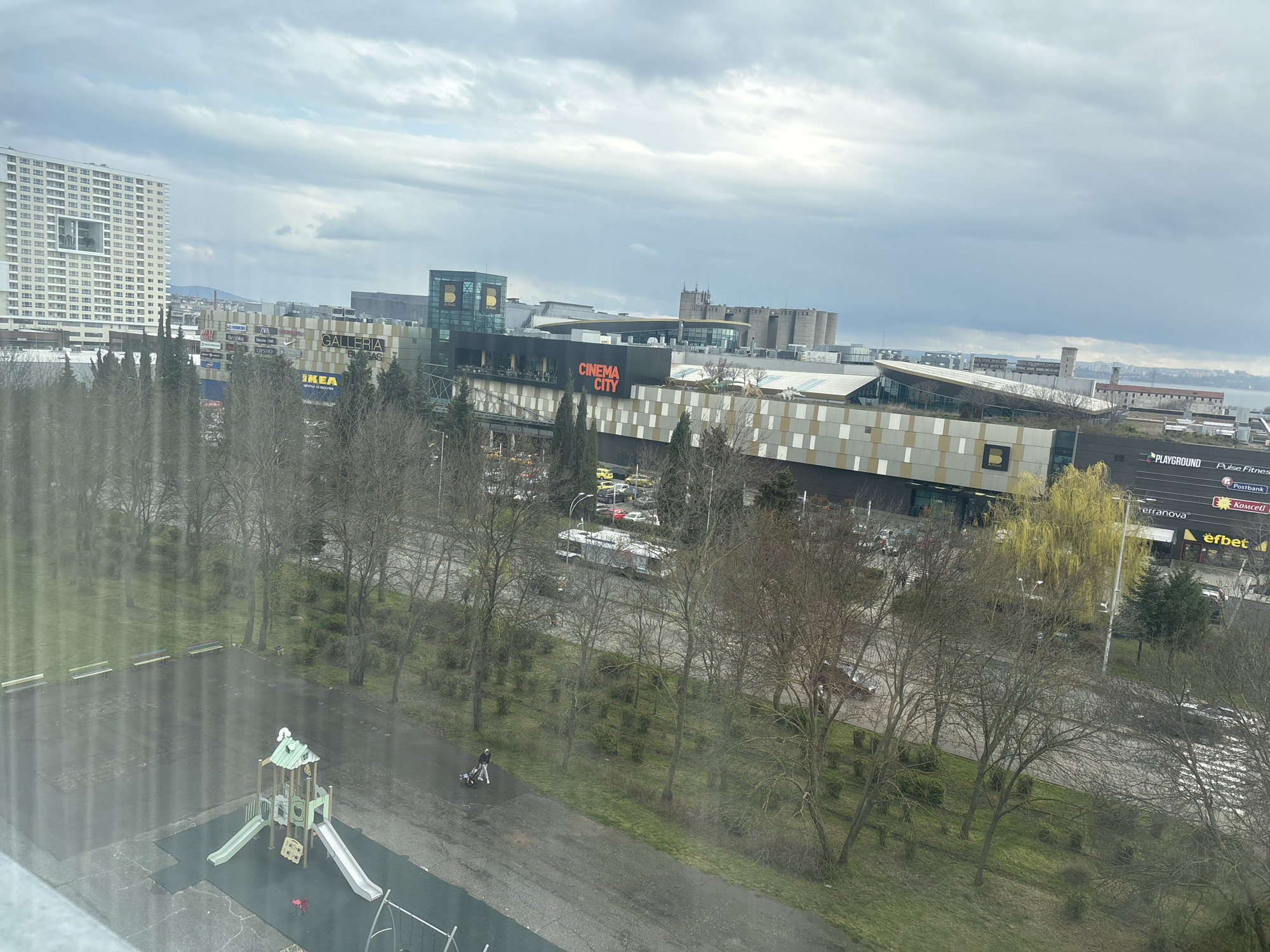 The image shows a view of part of Burgas, Bulgaria, from an elevated position. A large shopping complex featuring "CINEMA CITY" and other businesses like "IKEA" is visible. In the foreground, there's a children's playground area and a path where a person is walking, pushing a stroller. Trees with bare branches line the street. In the distance, buildings and other structures can be seen, and the sky is overcast.