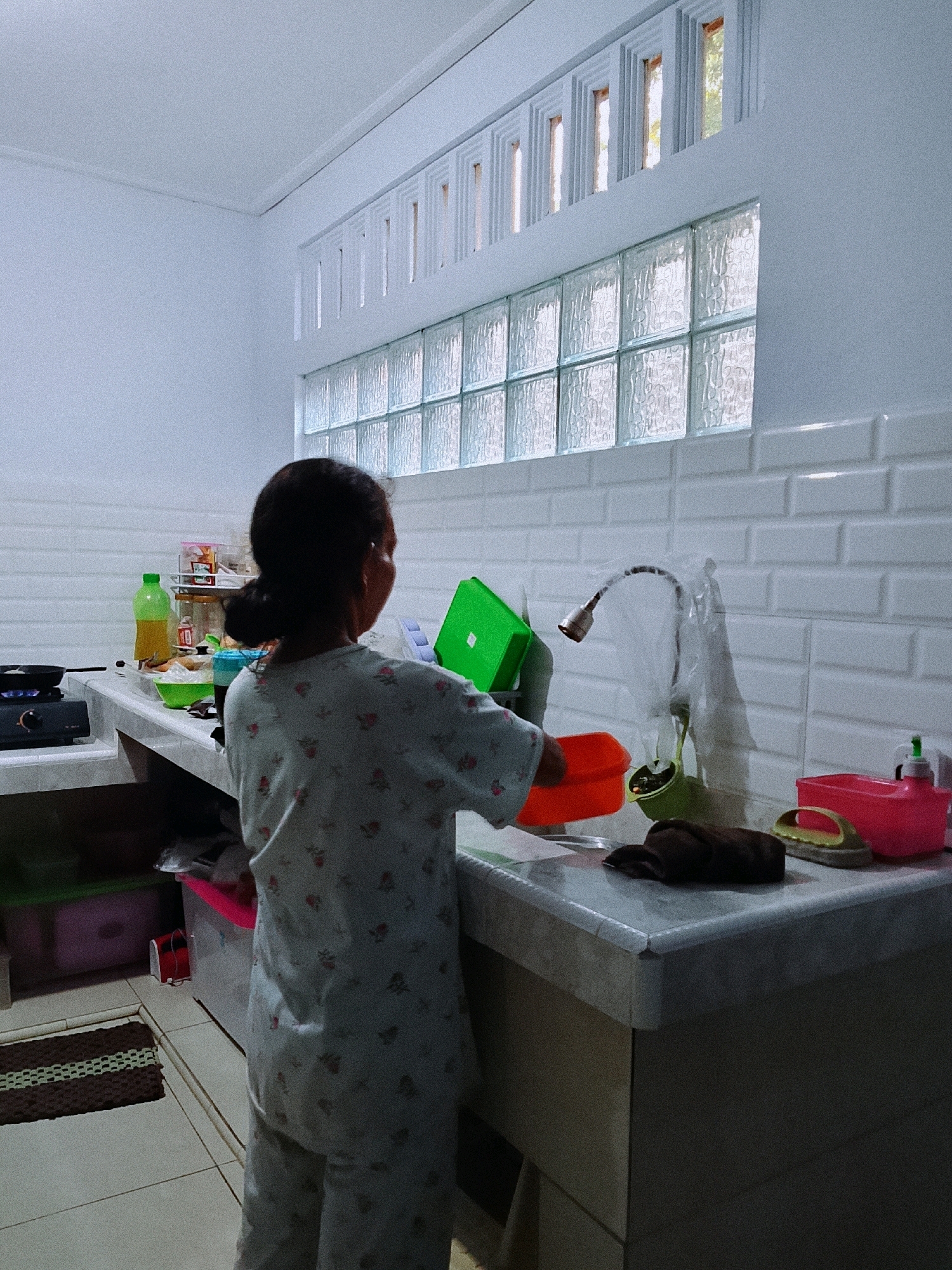 A person stands at a kitchen counter in what appears to be a residential kitchen in Kradenan, Indonesia. The person, seen from behind, is wearing a light-colored pajama set with a floral pattern and has dark hair pulled back. They are facing a sink area where various kitchen items are present, including a green plastic container, an orange basin, a faucet covered with a plastic bag, and a dish brush. To the left of the person, on the counter, is a gas stove with a pan on it, a green bottle of liquid, and other miscellaneous items. The kitchen walls are tiled with white subway tiles and a series of glass blocks form a window above the counter. The overall environment is illuminated by natural light coming from the windows and possibly indoor lighting, creating a slightly dim and muted atmosphere. The floor appears to be tiled and a dark mat with green stripes is visible near the bottom left of the image.
