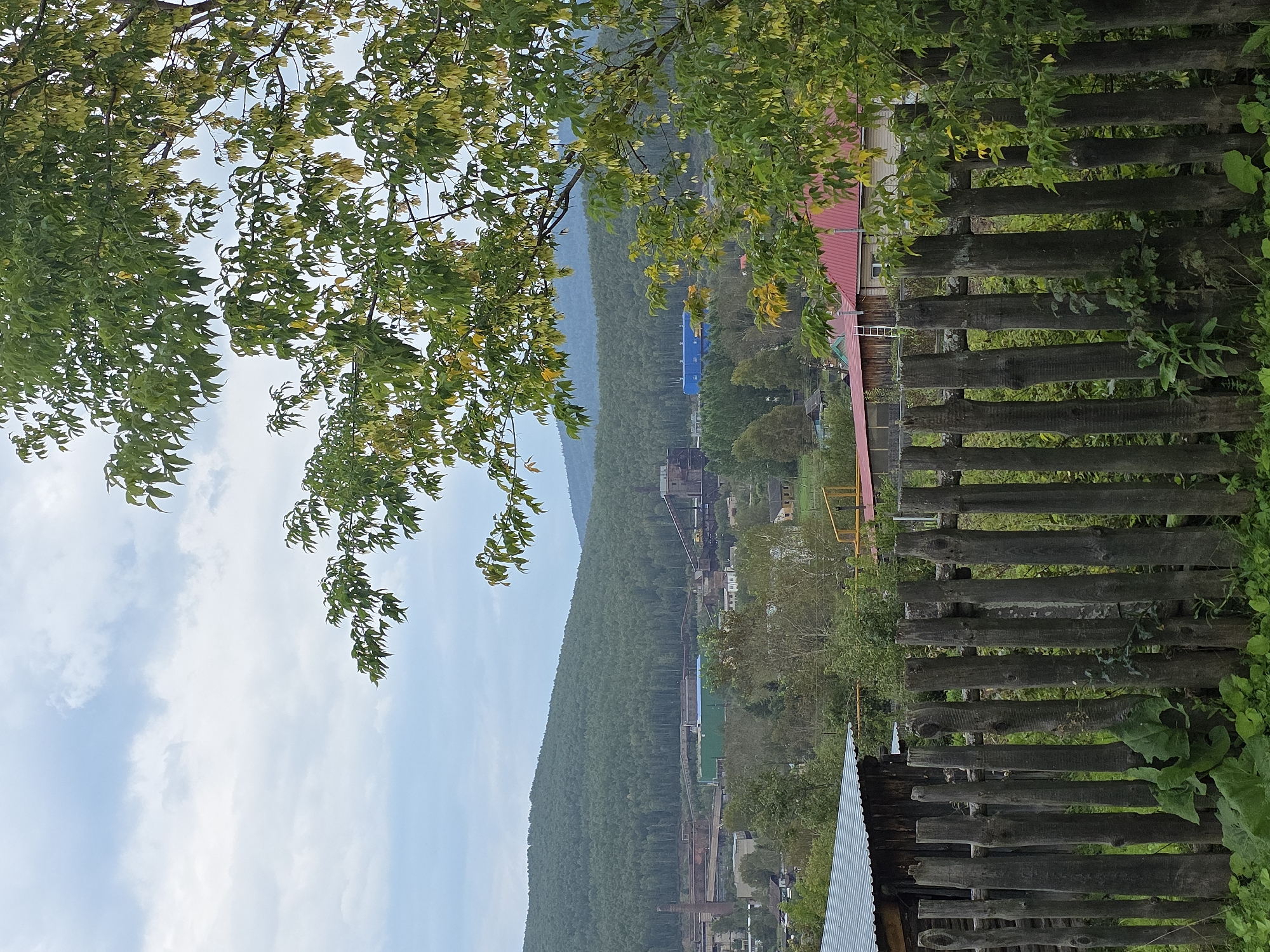 A weathered wooden fence runs across the foreground of the image, composed of roughly hewn vertical logs of varying widths. Behind the fence, a small cluster of rural buildings can be seen, some with corrugated metal roofs and others with red tiled roofs. A large tree with green and yellowing leaves partially obscures the view on the right side of the frame. In the midground, an industrial complex is visible, characterized by large factory buildings, smokestacks, and conveyor belts, set against a backdrop of densely forested hills. The sky above is overcast with scattered clouds. The scene appears to be located in the vicinity of the Magnitogorsk Iron and Steel Works in Magnitka, Russia, suggested by the industrial landscape and surrounding natural environment. There are no people or discernible actions in the image.