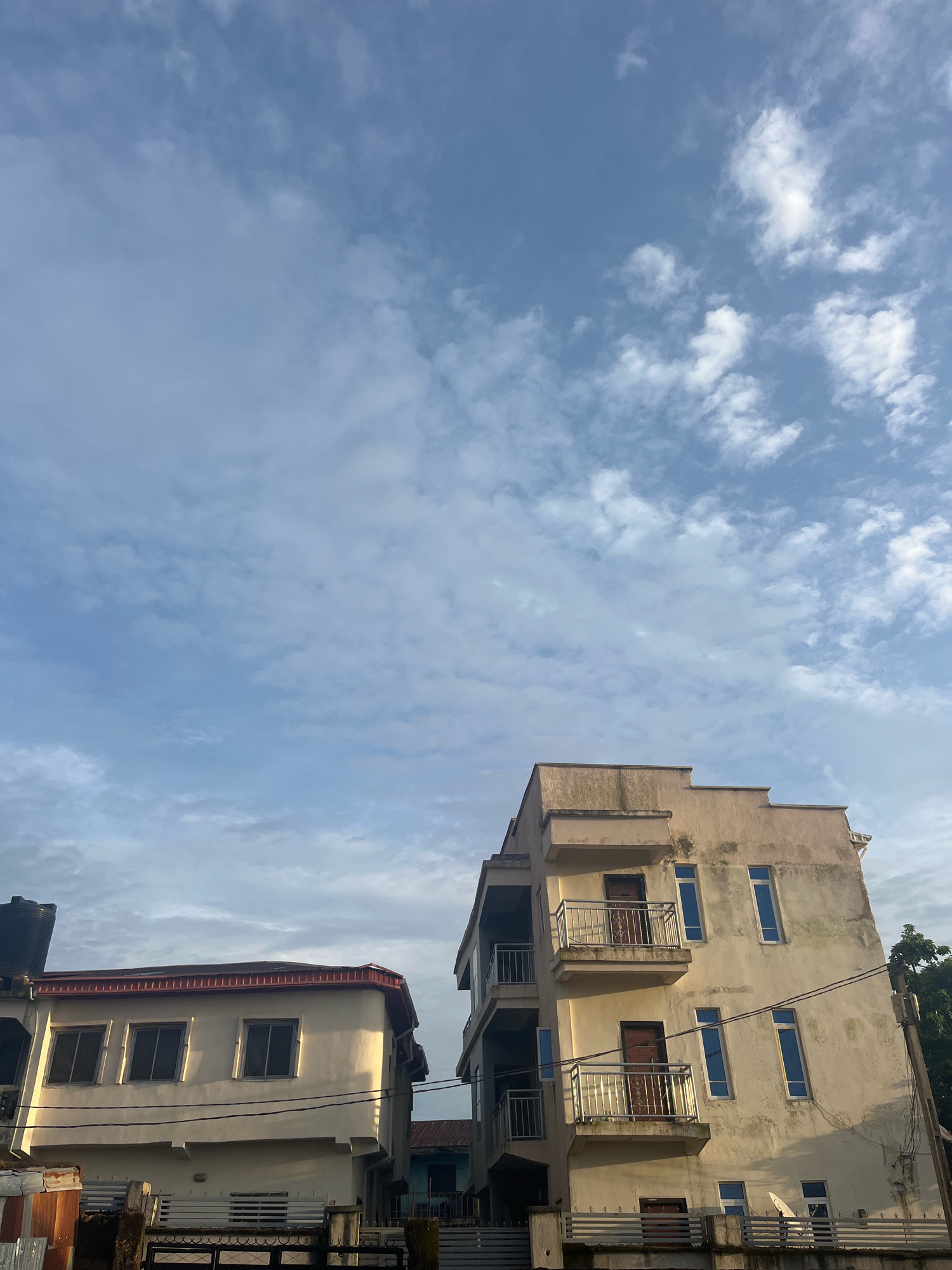 The image shows a street view in Jos, Nigeria, on a clear day with scattered clouds. In the foreground, two multi-story buildings are visible. The building on the left is lighter in color with a red-trimmed roof and several windows on its facade. A black water tank is mounted on top of this building. To its right, a taller, cream-colored building features multiple balconies with metal railings and several tall, narrow windows. The street in front of the buildings appears to have fencing and some debris. The sky above is a light blue with white, wispy clouds.
