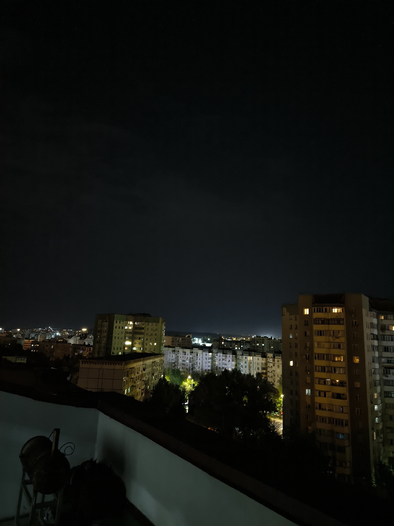An elevated, nighttime outdoor shot captures a cityscape with a dark sky dotted with faint stars. In the foreground, a dark-colored terrace railing and a portion of a balcony wall are visible. To the left on the balcony, two dark, vaguely cylindrical objects with metal framing and handles are present.
The middle ground and background reveal a dense urban environment with multiple apartment buildings. To the right, a tall, light-brown apartment building with numerous lit windows is prominent. Adjacent to it, to the left, is another multi-story building, partially obscured by trees, also showing illuminated windows. Further to the left, more apartment buildings with lit windows extend into the distance, forming a sprawling city panorama under the night sky. The scene is characteristic of a residential area in Chișinău, Moldova, likely captured from an apartment building's balcony.