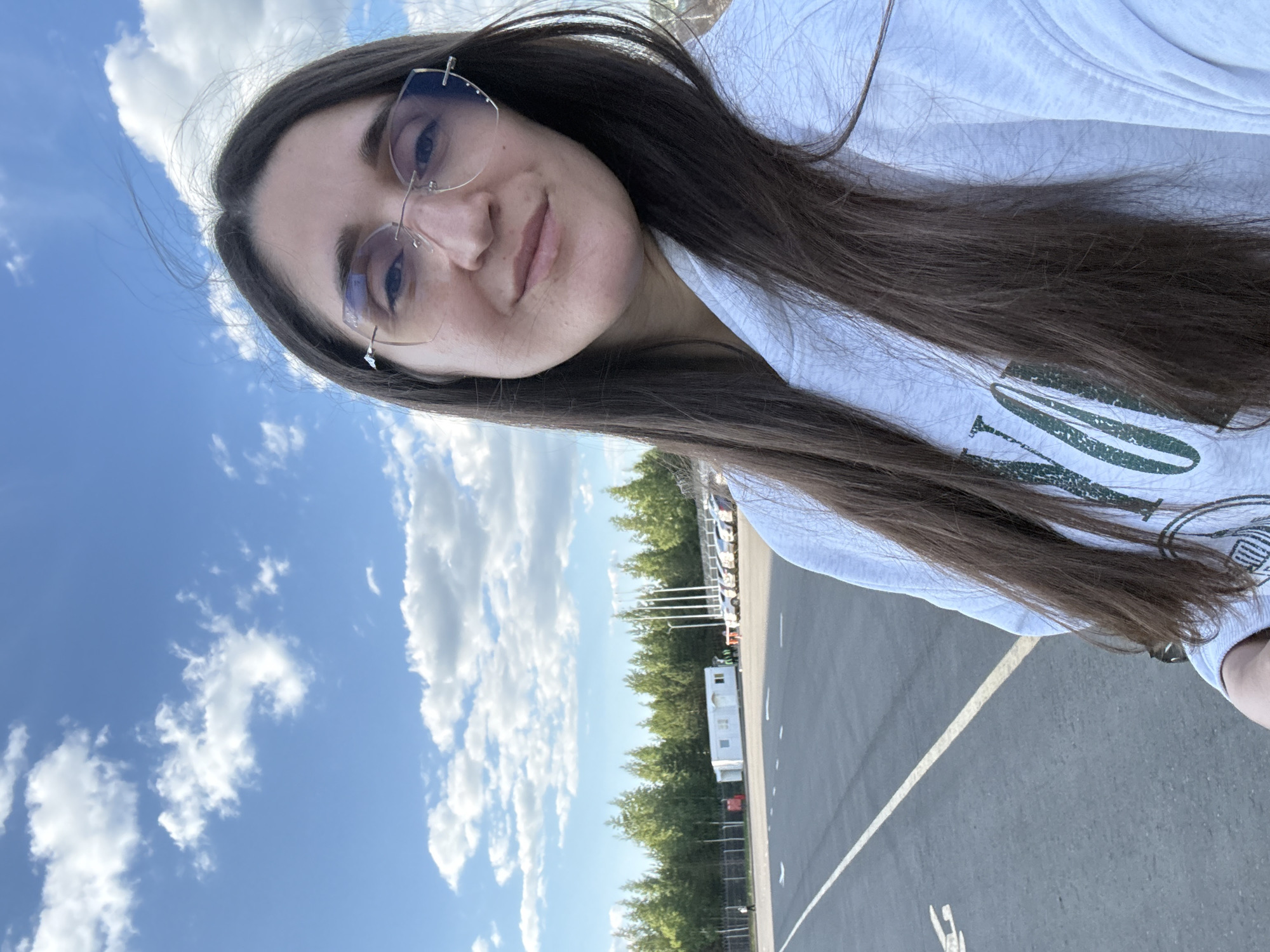 A forensic examination of the image reveals a detailed scene in Sarapul, Russia, featuring a woman taking a selfie outdoors on a bright day. The subject, positioned in the foreground and slightly right of center, is a woman with long, dark brown hair parted in the middle. She wears rimless glasses with light-tinted lenses and a light-colored (likely white or light grey) hooded sweatshirt featuring a partially visible green graphic on the chest. She is looking directly at the camera with a subtle smile.
The immediate environment consists of a vast expanse of dark grey asphalt or concrete pavement, typical of an airfield, test track, or large parking area. Distinct white lines and symbols are painted on the pavement, including a prominent solid white line extending diagonally from the lower left towards the upper right, and faded arrow-like markings.
In the midground, towards the left, a solitary individual is visible walking along a fence line. Further back, a white, rectangular modular building or container with a pitched roof stands prominently. To the right of this structure, a metal fence separates the paved area from a row of light-colored parked vehicles, possibly cars, which are partially obscured. Several tall, slender light poles rise behind the parked vehicles.
The background features a dense line of conifer trees, forming a natural barrier or forest line beyond the operational area. Above, the sky is clear blue with numerous scattered white cumulus clouds. The overall lighting suggests a sunny daytime setting.
