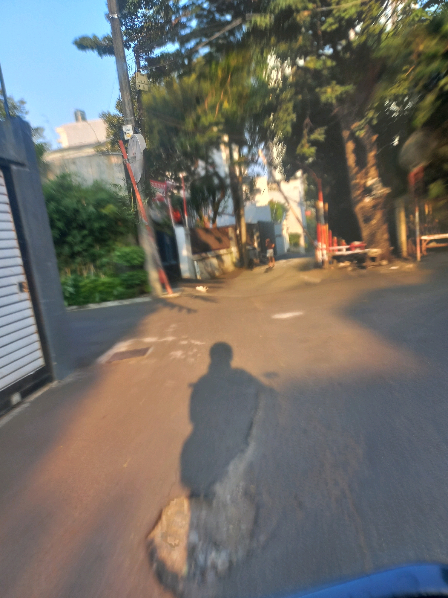 The image depicts a street scene in South Jakarta, Indonesia, characterized by a blurred, impressionistic quality. A prominent feature is the shadow of a person, likely the photographer, cast on the asphalt road, indicating the sun is low in the sky, suggesting either early morning or late afternoon. The road surface appears worn in places, with a visible patch of rougher material. To the left, a portion of a grey, textured wall and a white garage door with horizontal slats are visible. Beyond this, lush green foliage can be seen. In the background, buildings and trees are out of focus, but one can discern elements of urban infrastructure, including a utility pole with various attachments, including a satellite dish. The presence of red and white striped poles and barriers suggests a controlled access point or a construction area. In the middle ground, a solitary figure stands on the road, also out of focus. The overall atmosphere is one of a typical urban environment in South Jakarta, bathed in the warm light of the setting or rising sun.
