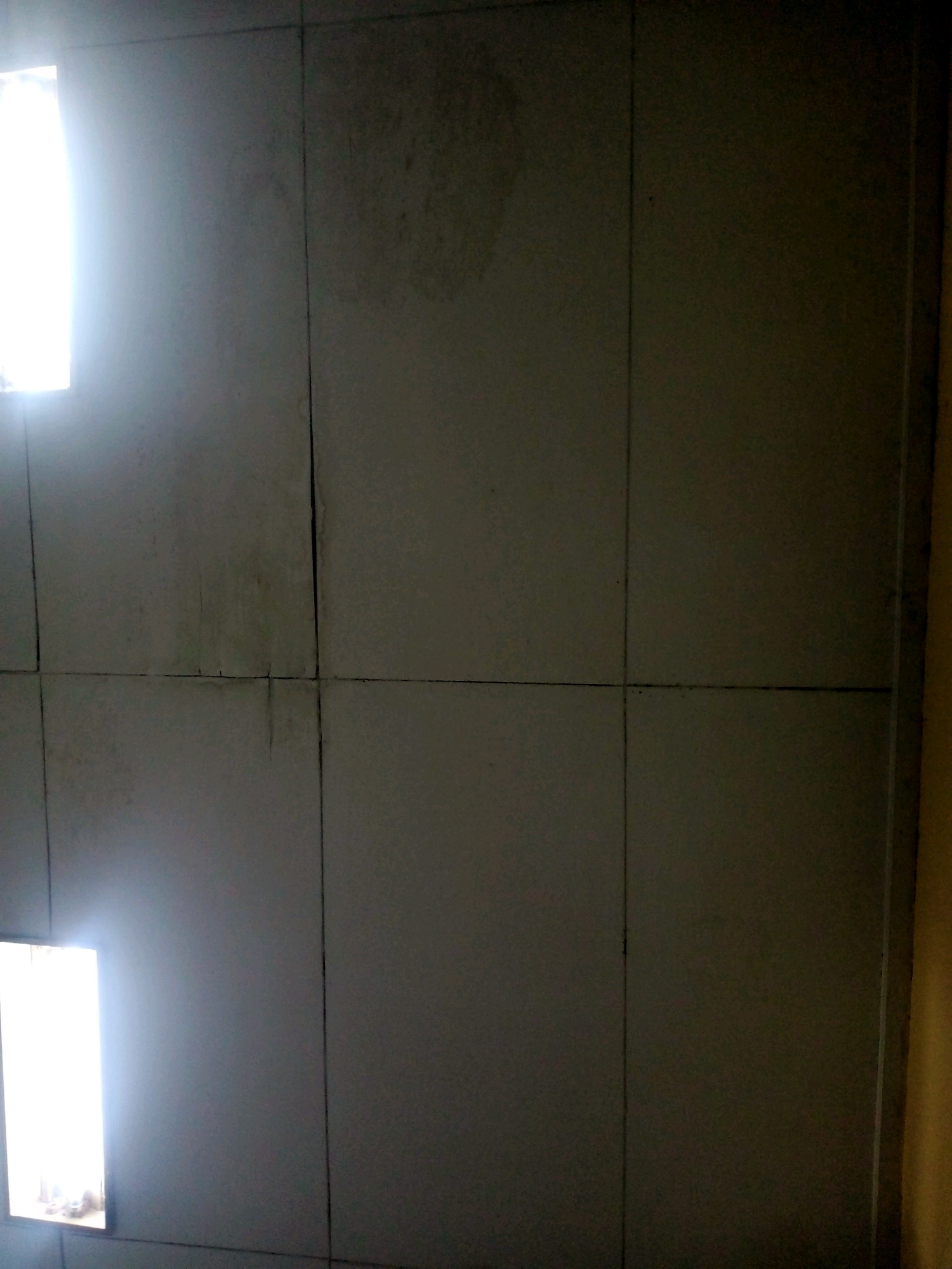 The image depicts a close-up view of a wall, possibly an interior wall, divided into large square tiles. The tiles are a dull, grayish-white color and appear somewhat aged, with visible grout lines forming a grid. In the upper left quadrant, a bright light source, possibly a window or a light fixture, illuminates a portion of the wall, creating a strong glare and obscuring details in that area. There are some faint discolorations or stains on the tiles, particularly in the upper half of the image, suggesting water damage or aging. The lower half of the image is darker and shows more of the tiled surface. The overall impression is one of a utilitarian or perhaps neglected surface. No people, specific objects, or actions are clearly identifiable in the frame. The context of Cangkuang Wetan, Indonesia, does not provide any specific visual cues within the image itself.